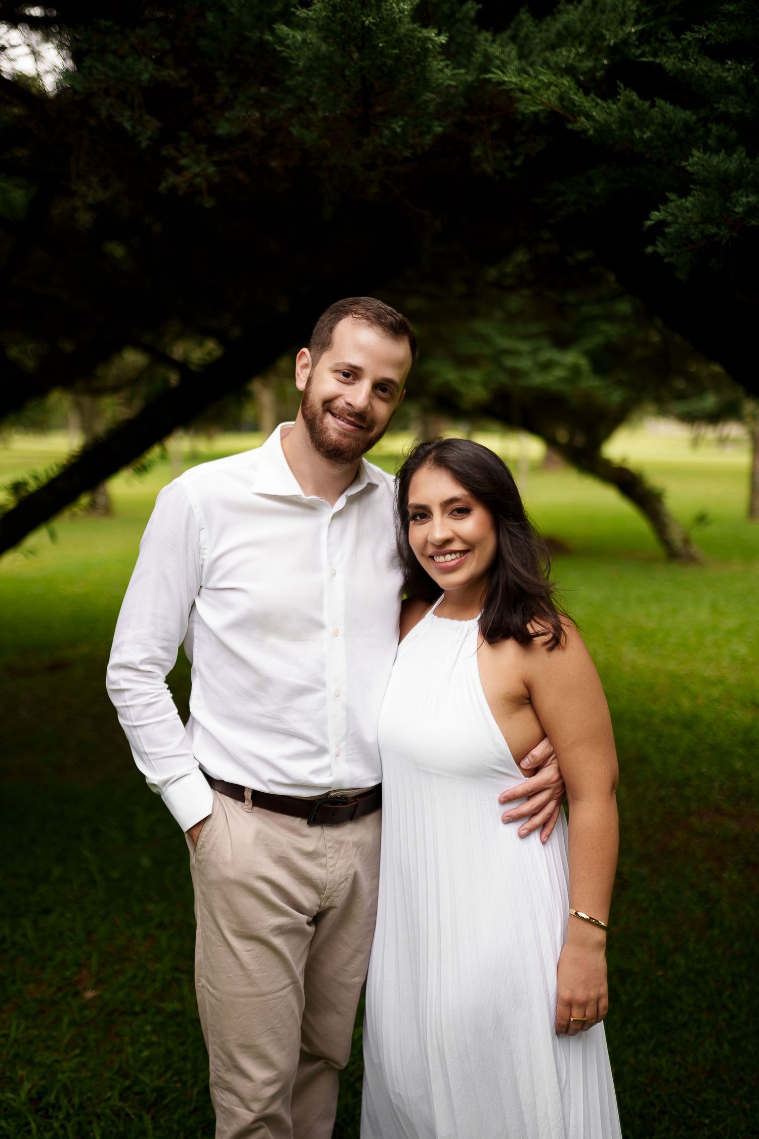 Smiling couple in white outfits posing together in a lush green park with large trees in the background.