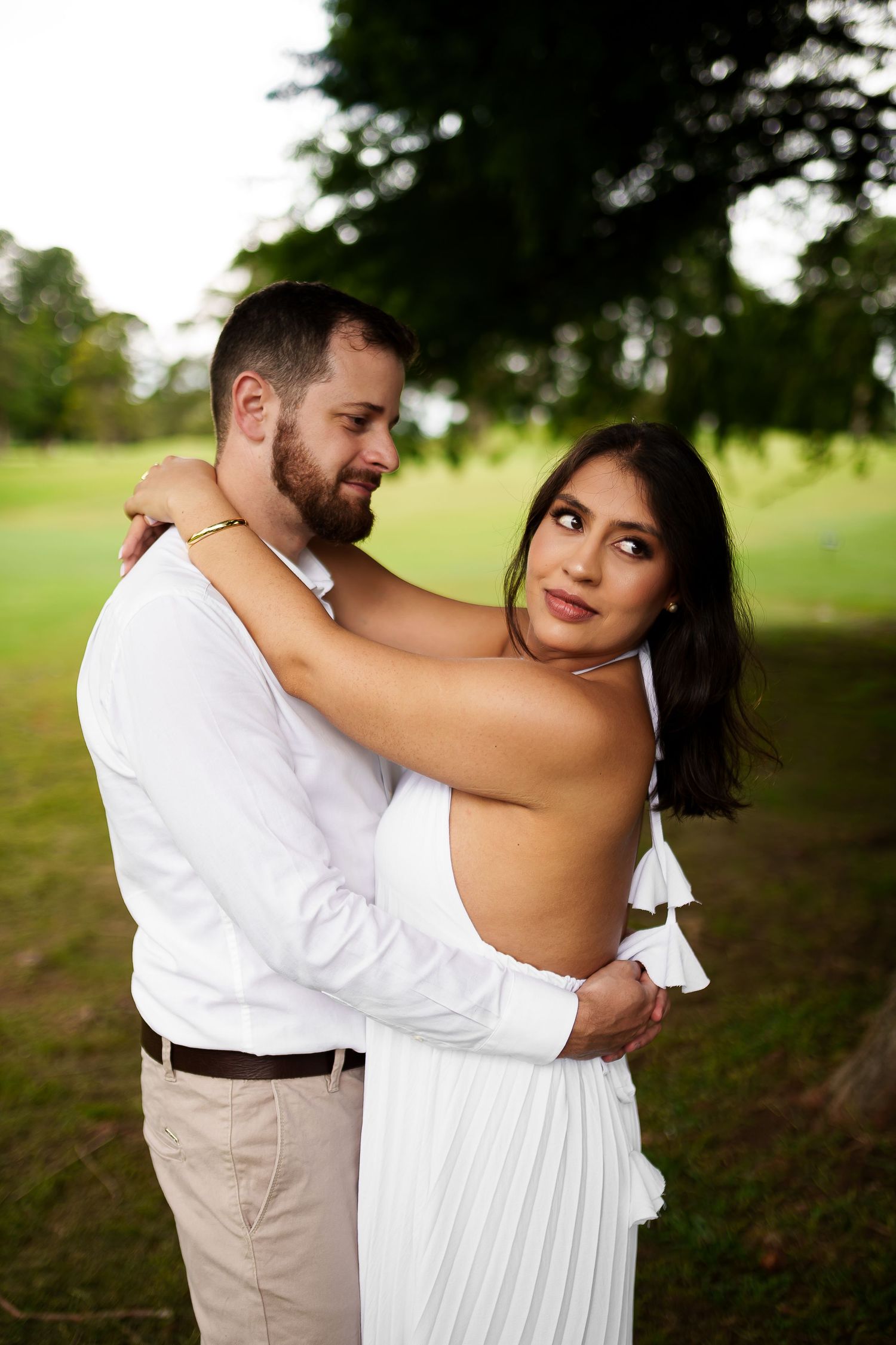 Couple in white outfits embracing outdoors, woman looking at camera, lush green park background, romantic portrait.