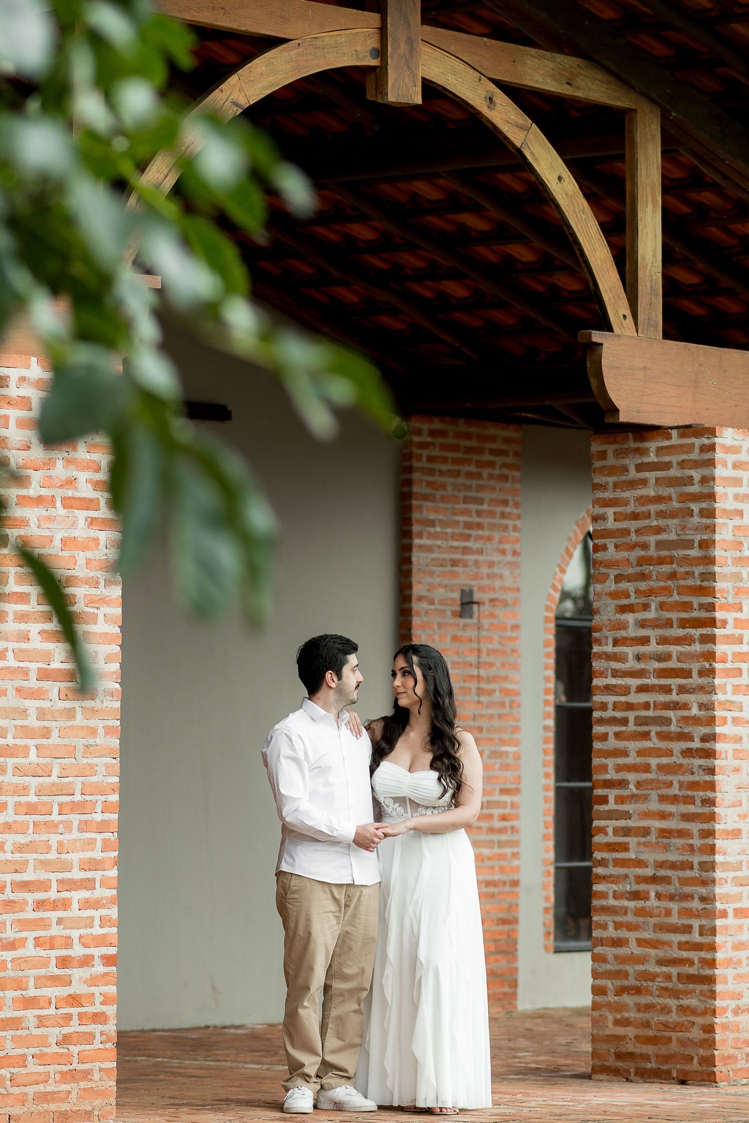 Couple in white outfits holding hands under a rustic brick archway with wooden beams at an outdoor venue.