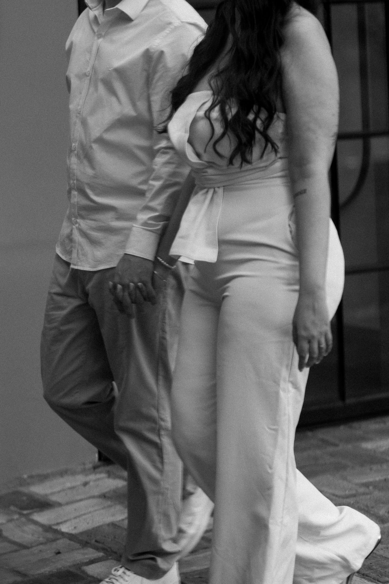 Black and white photo of a couple holding hands, woman in white pantsuit and man in button-down shirt leaning against a wall.