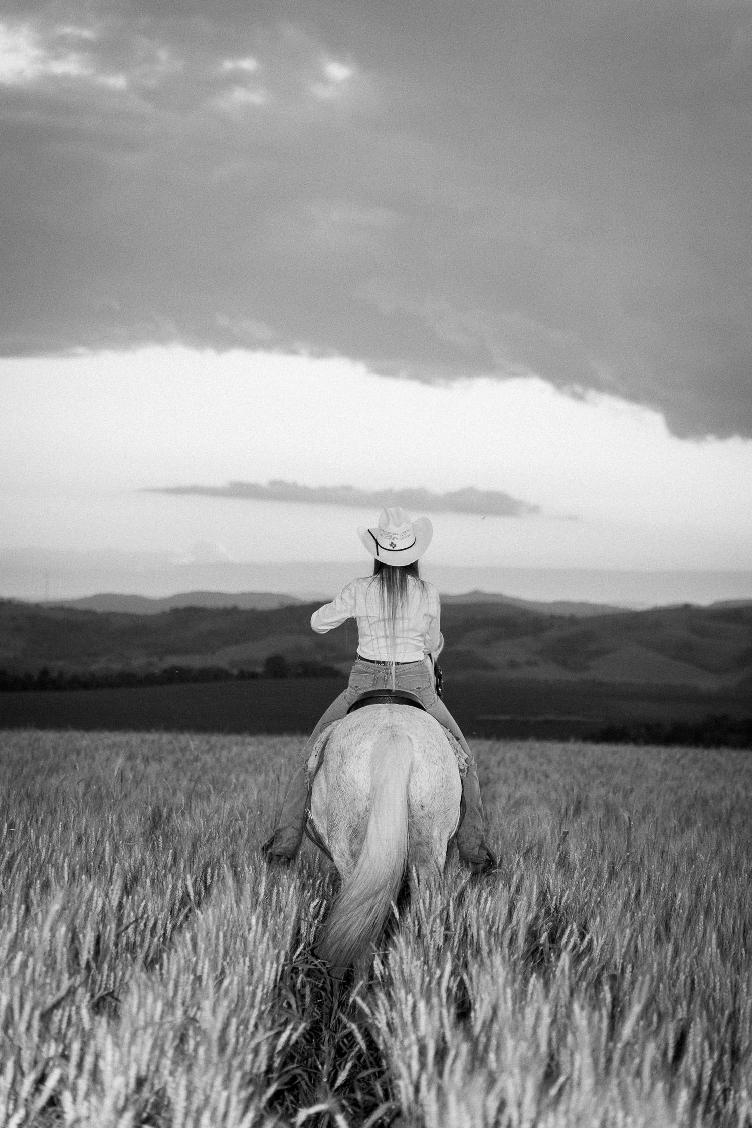 Black and white photo of a woman in cowboy hat riding a horse through a wheat field with dramatic stormy sky.