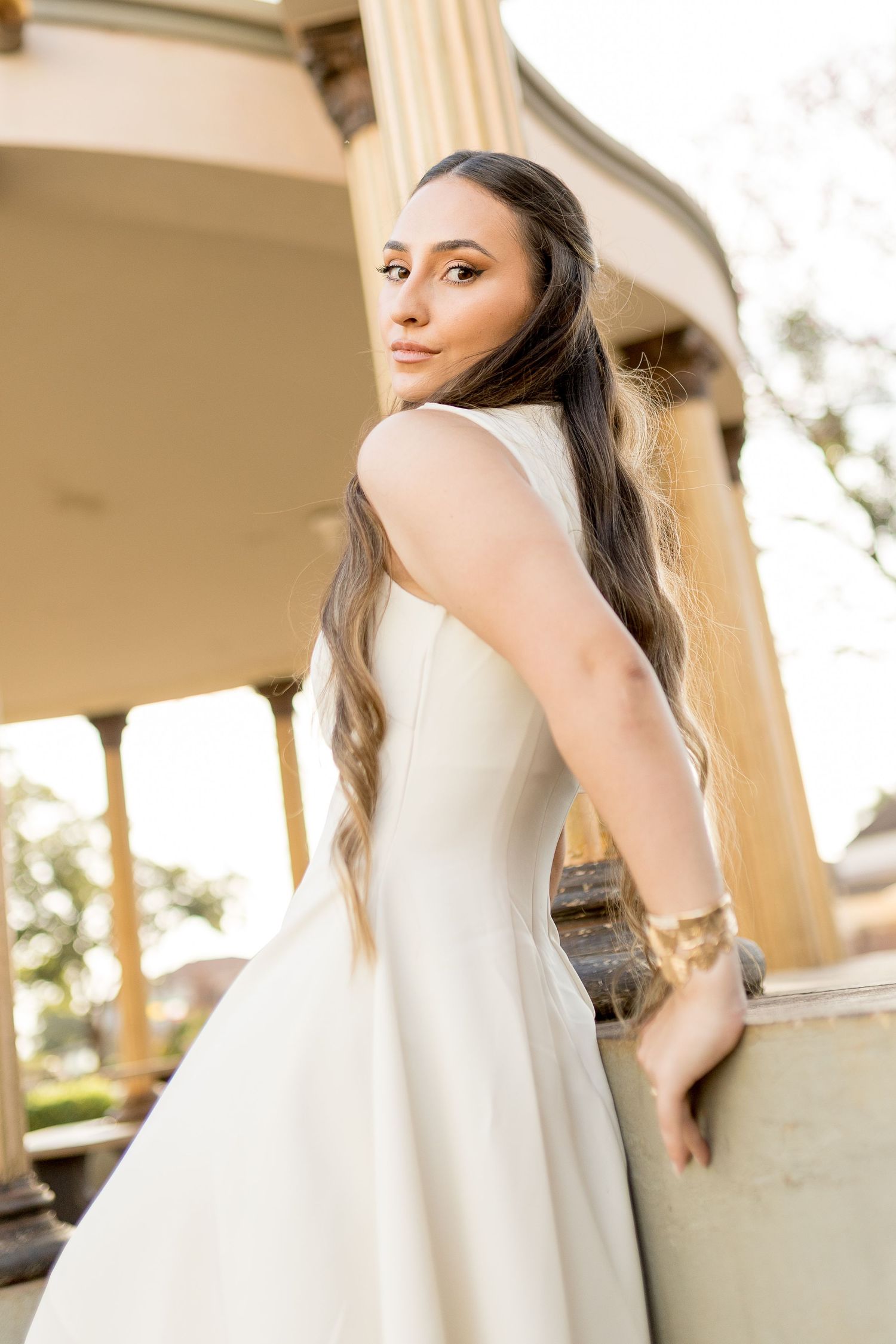 A bride in a white sleeveless gown poses gracefully by a column, her long hair flowing, wearing a gold bracelet.