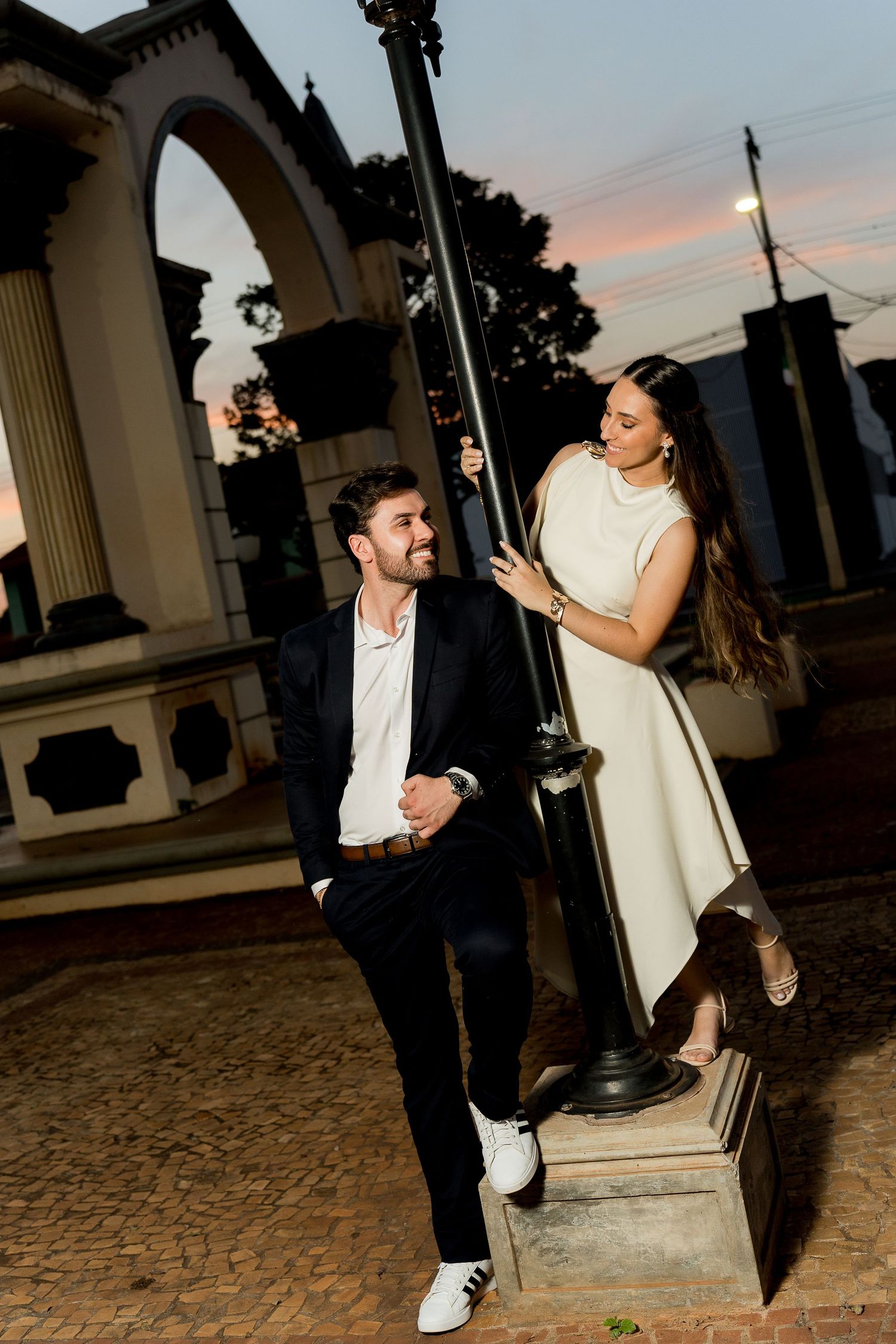 A couple poses by a lamp post at dusk, man in black suit, woman in white dress, with an arch structure behind them.