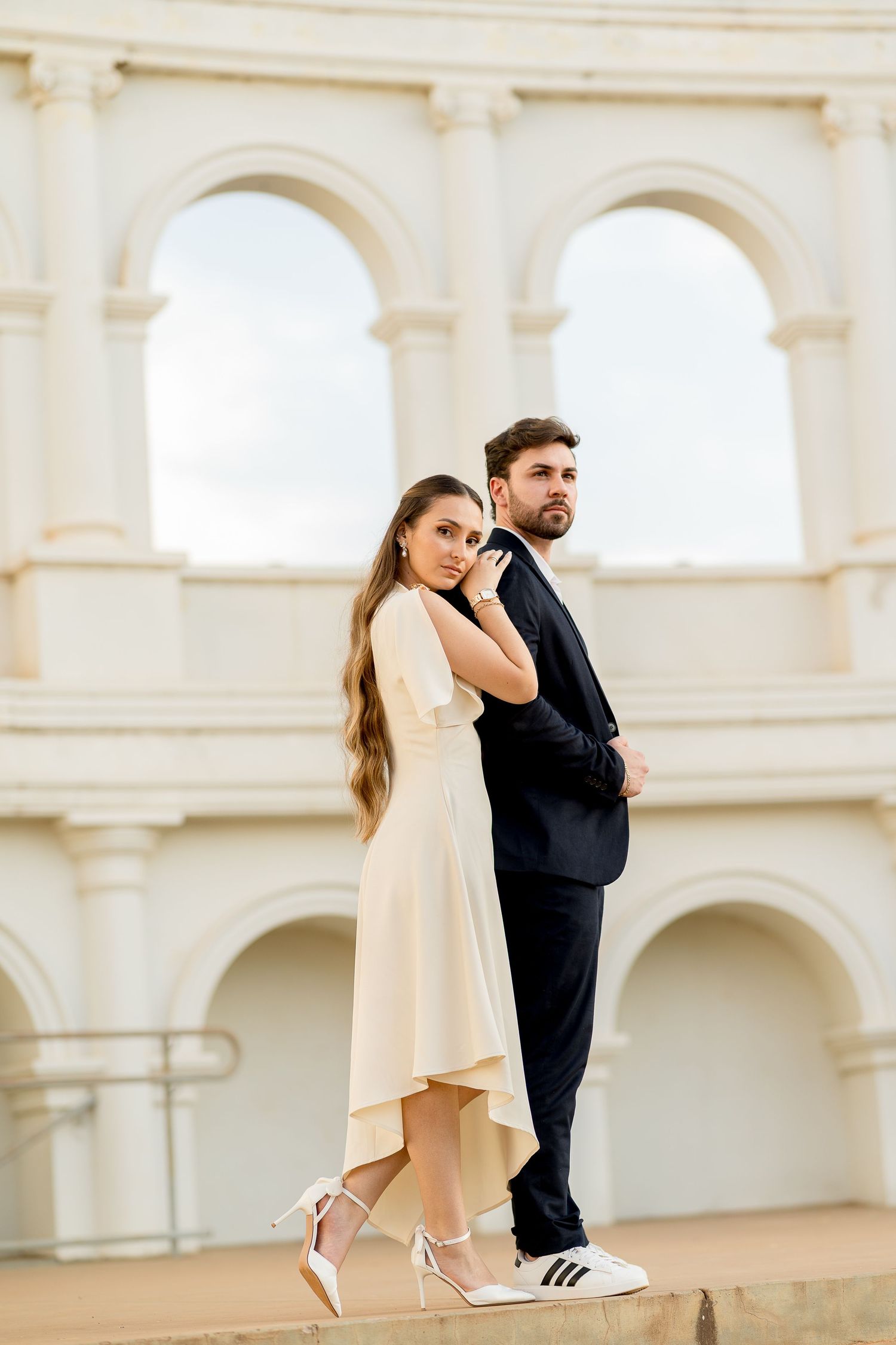 Elegant couple posing in front of classical arched building, woman in cream dress, man in dark suit.