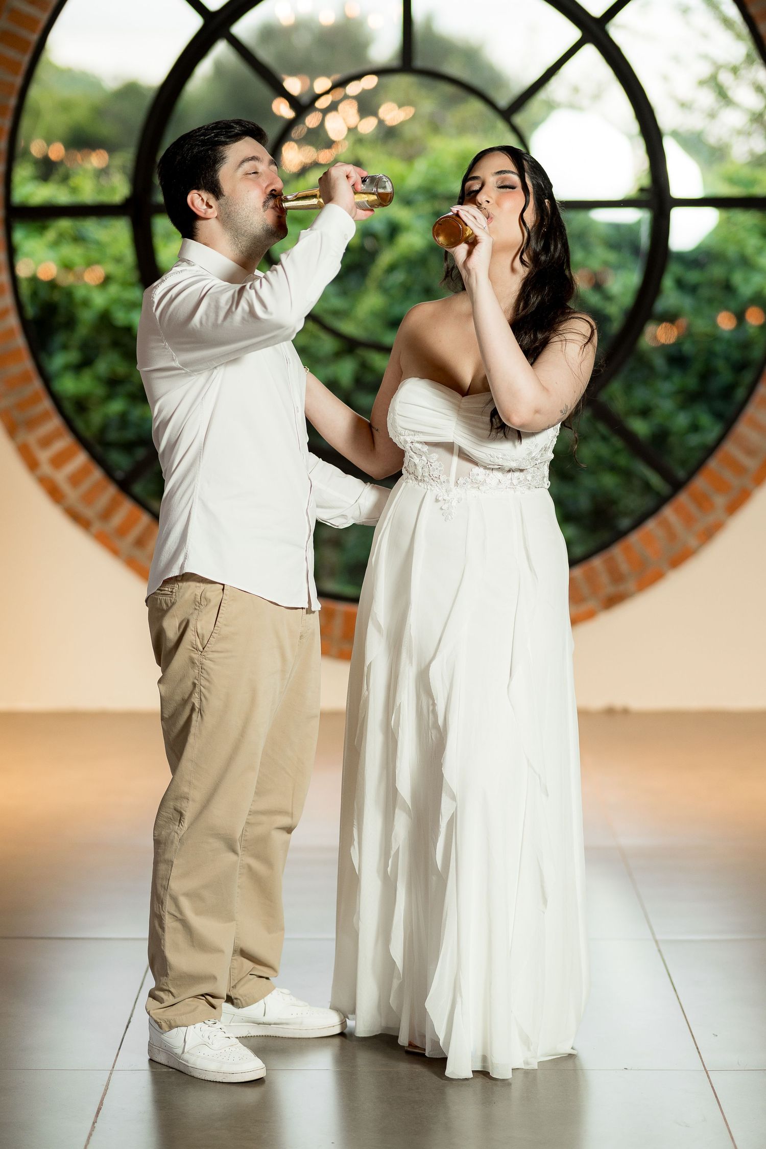 Bride in white gown and groom in white shirt toast with drinks before circular window at wedding reception.