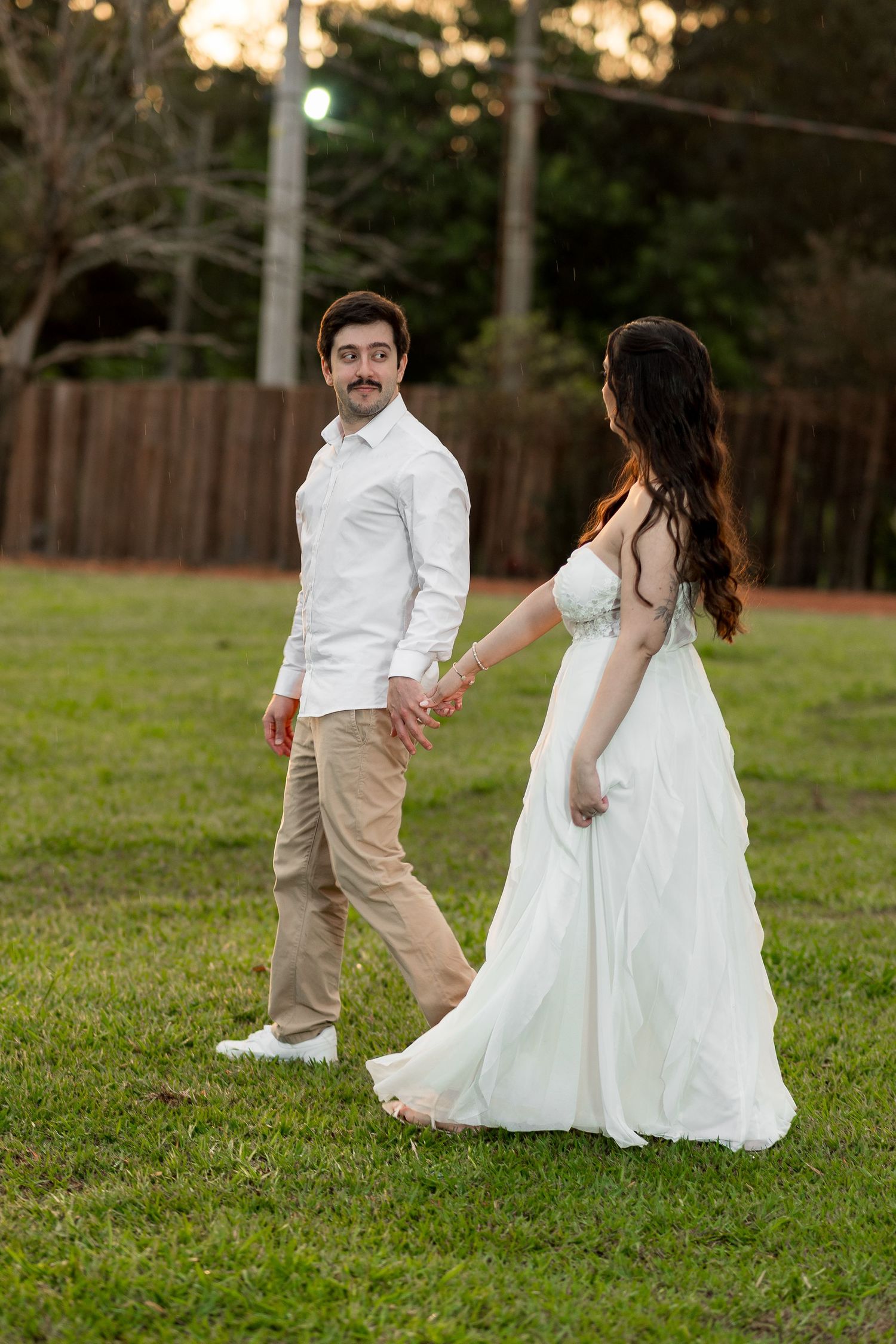 Couple in wedding attire holding hands walking on green grass, groom in white shirt looking back at bride in flowing white gown.