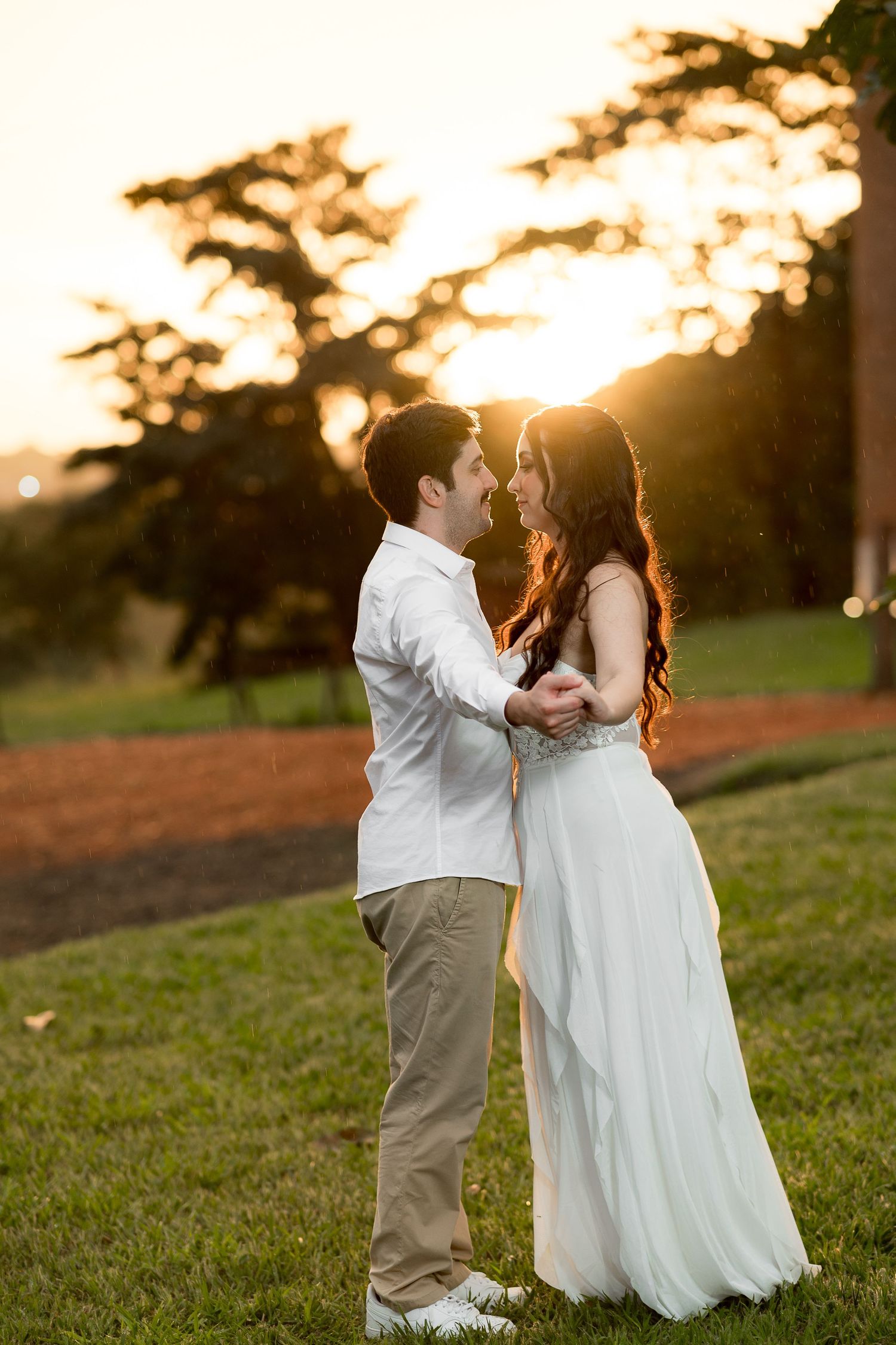 Couple sharing a romantic dance at golden hour sunset on green grass, woman in flowing white gown, man in white shirt.