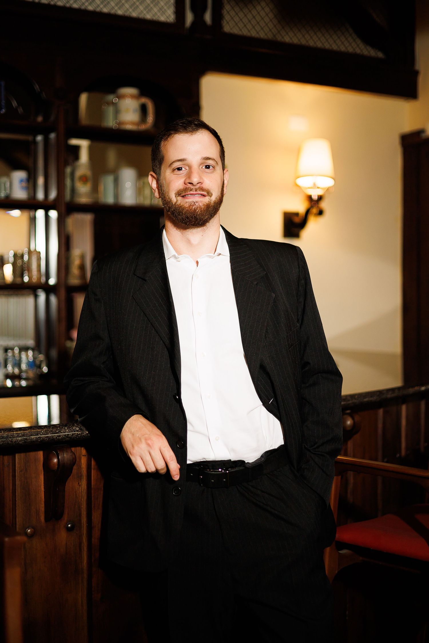 Bearded man in black pinstripe suit and white shirt leaning against a bar in an elegant restaurant.