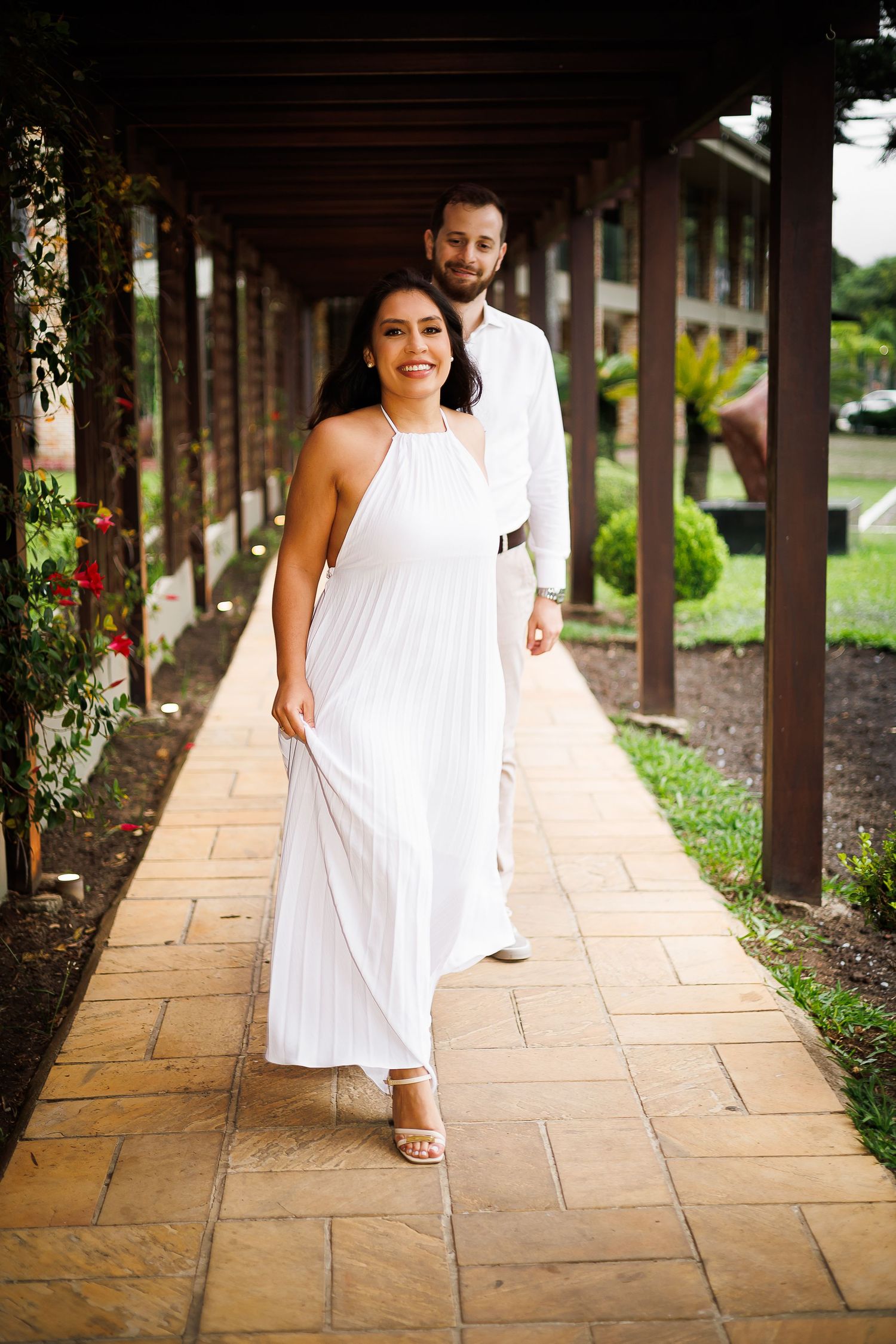 Couple in white outfits walking along a covered garden pathway, woman in halter maxi dress smiling.