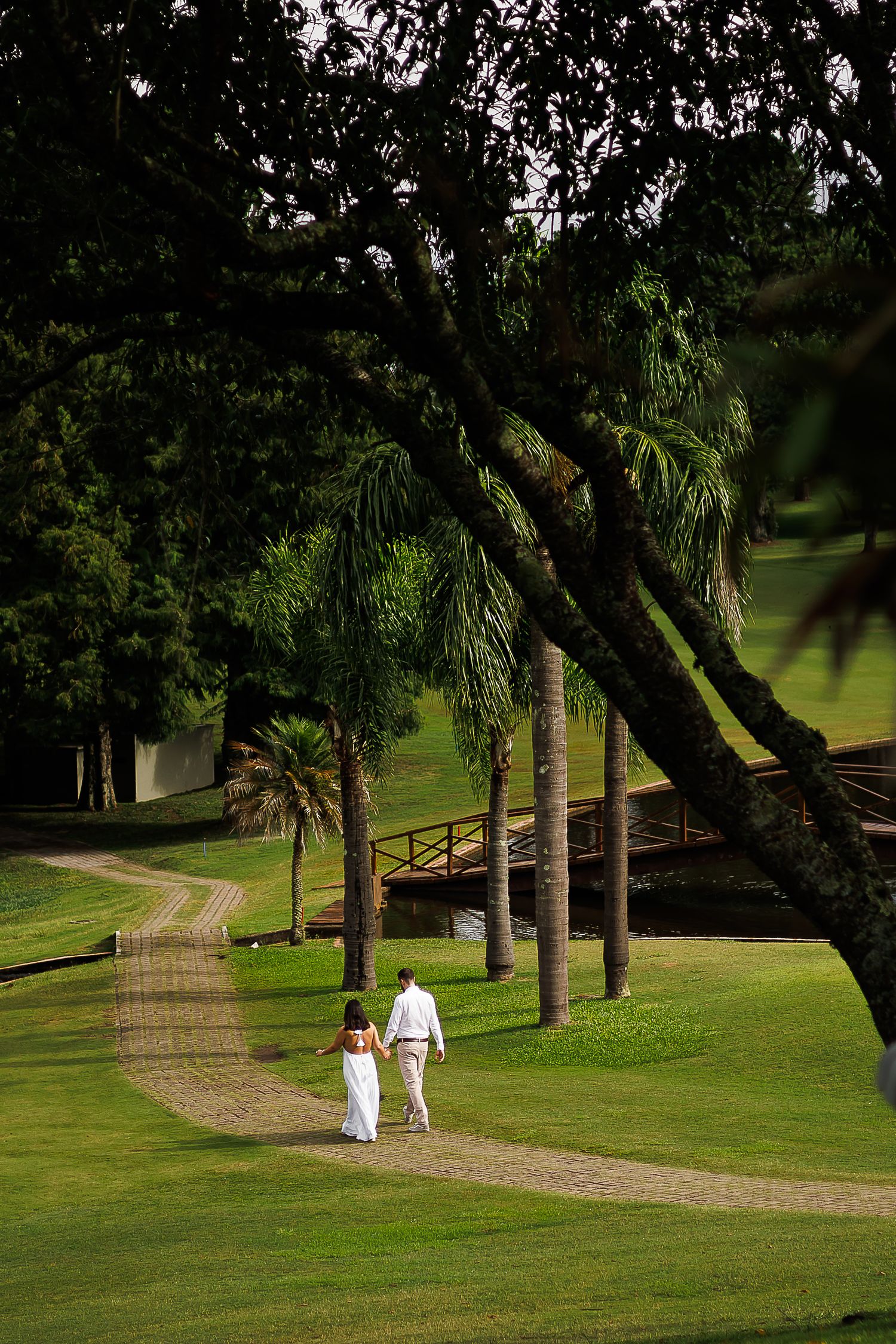 A couple in white attire walks hand-in-hand along a brick path through a lush green park with palm trees.