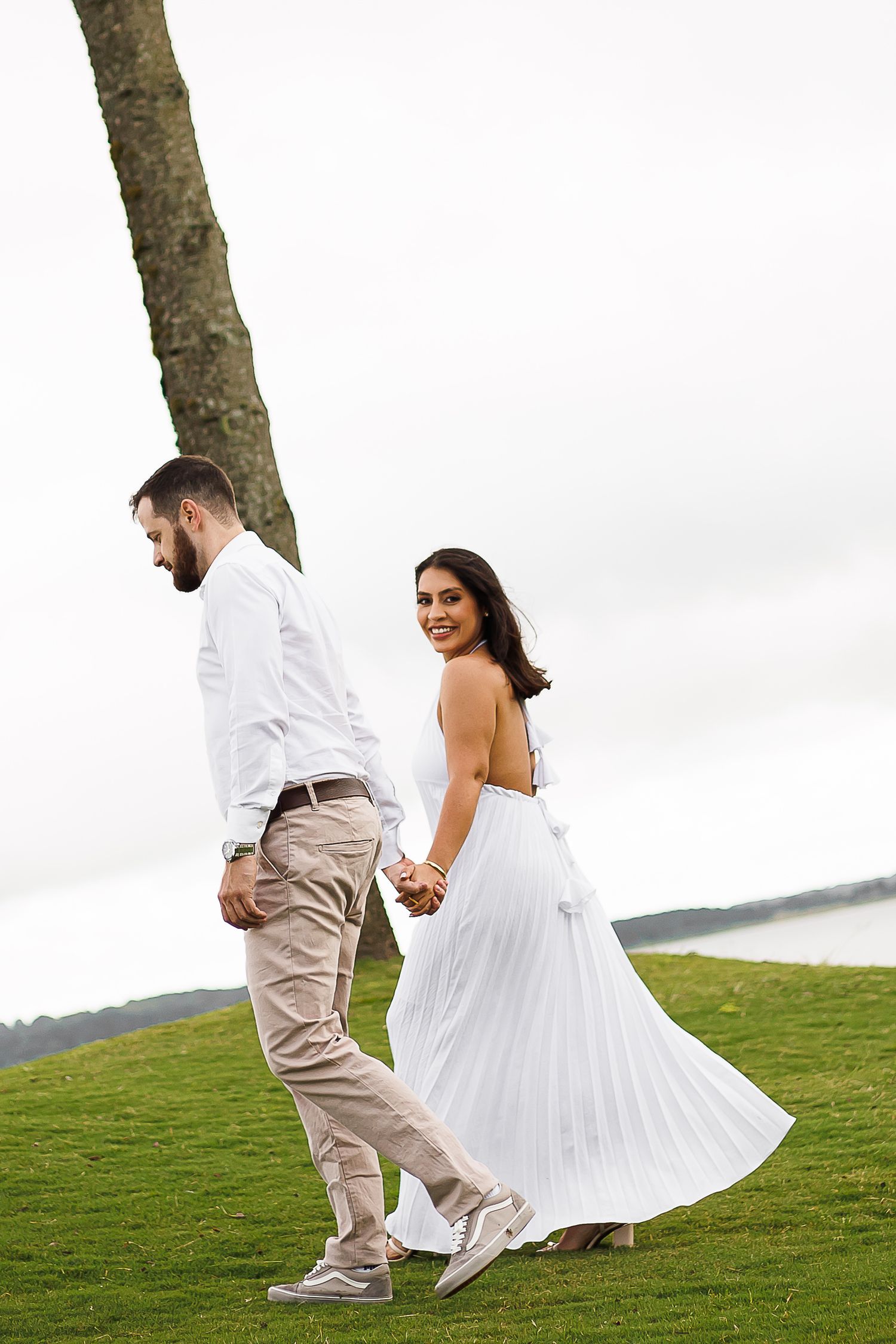 Couple in white attire holding hands walking on green grass near a tree with water in the background.