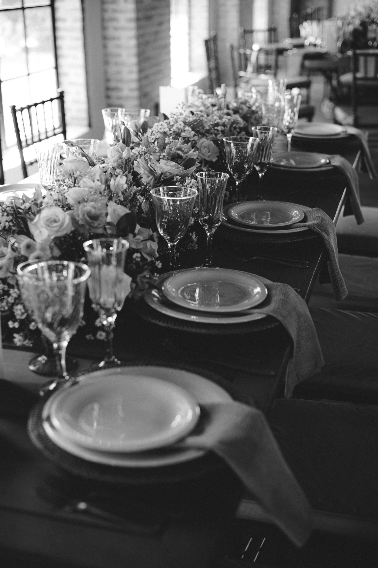 Black and white photo of elegant wedding reception table with floral centerpiece, crystal glasses, and layered place settings.