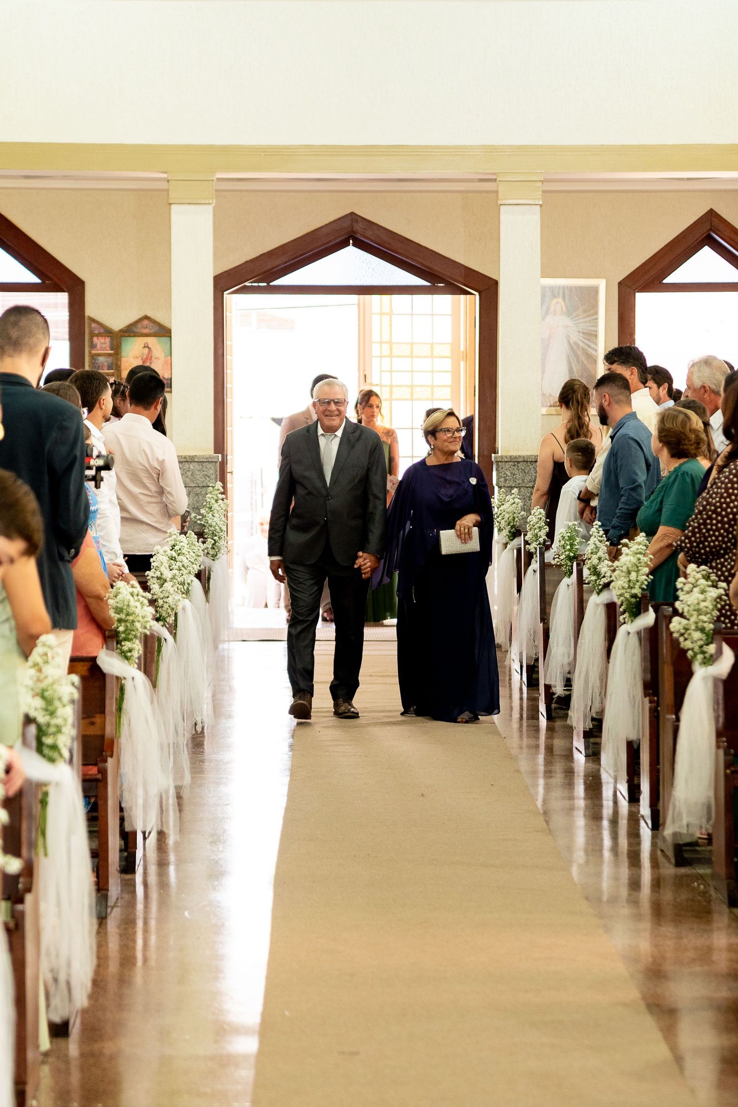 Couple walks down church aisle at wedding ceremony, pews decorated with white flowers and tulle.