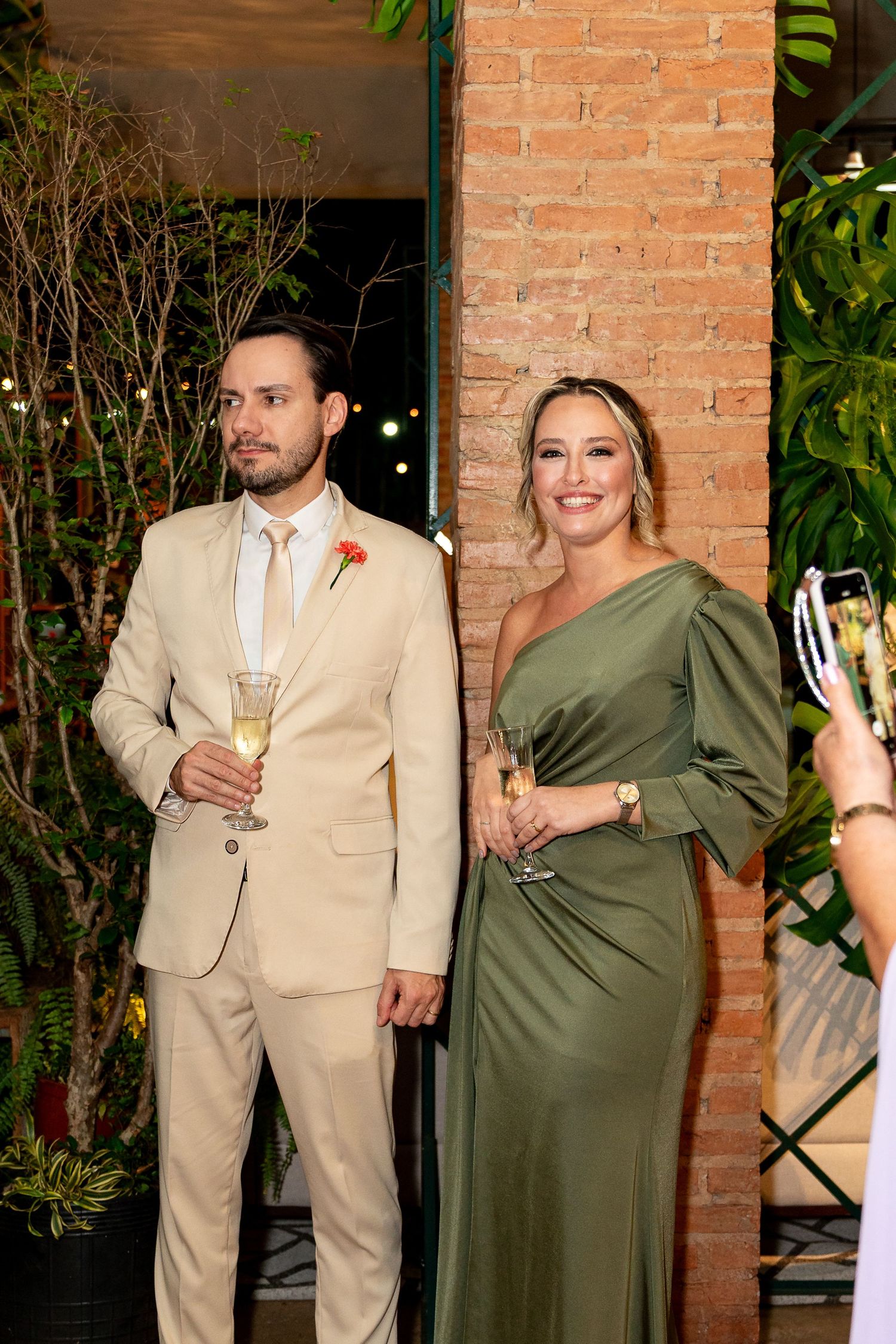 Elegant couple in formal attire holding champagne glasses at an event, man in beige suit, woman in olive green gown.