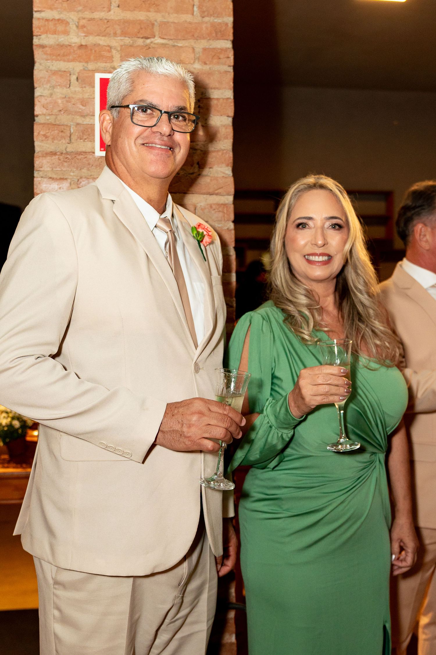 Elegant couple at a wedding reception, man in beige suit with boutonniere, woman in green dress, both holding champagne glasses.