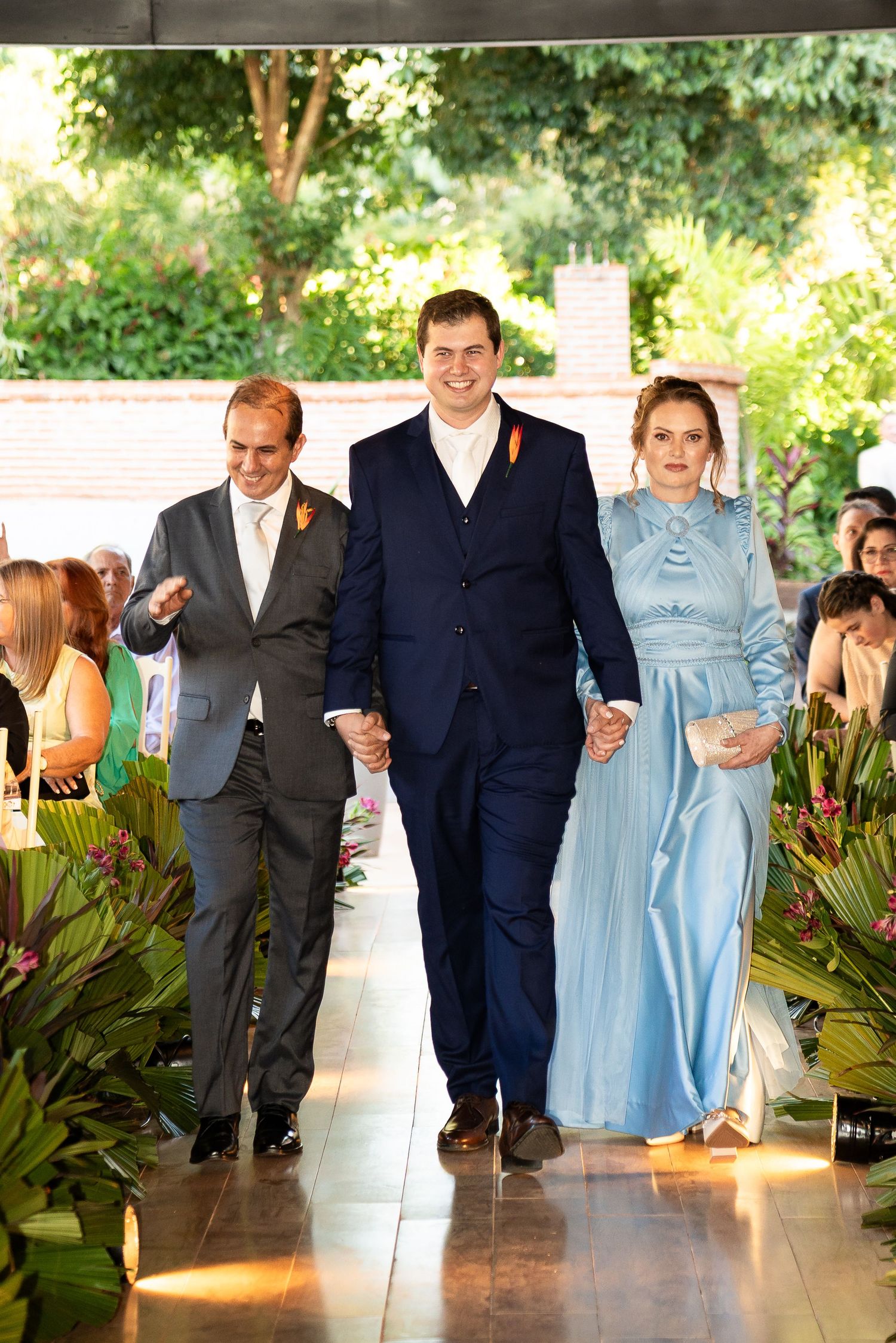 Groom walks down wedding aisle flanked by parents, wearing navy suit, as guests watch in lush green venue.
