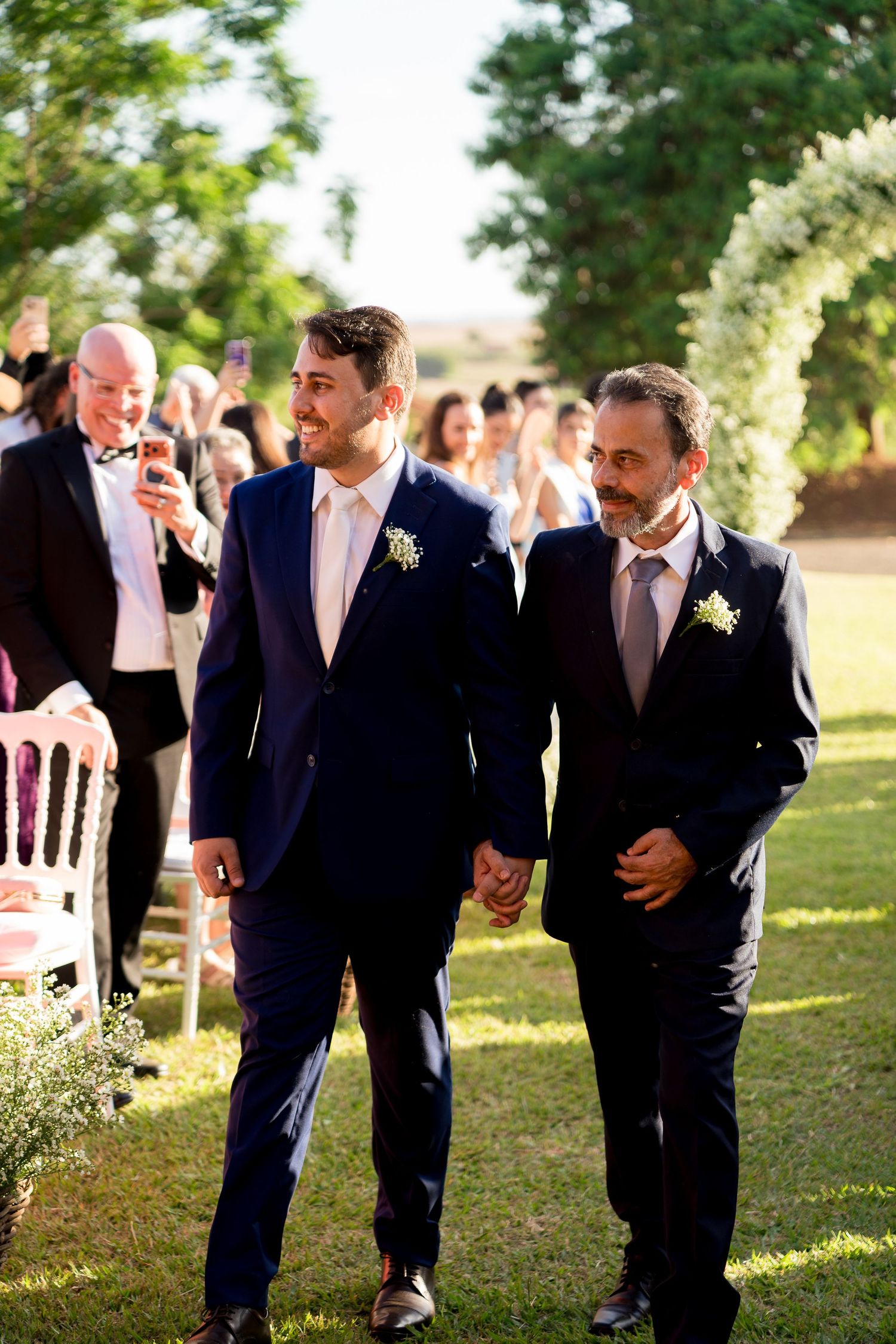 Two grooms in navy suits hold hands while walking down the aisle at an outdoor wedding ceremony.