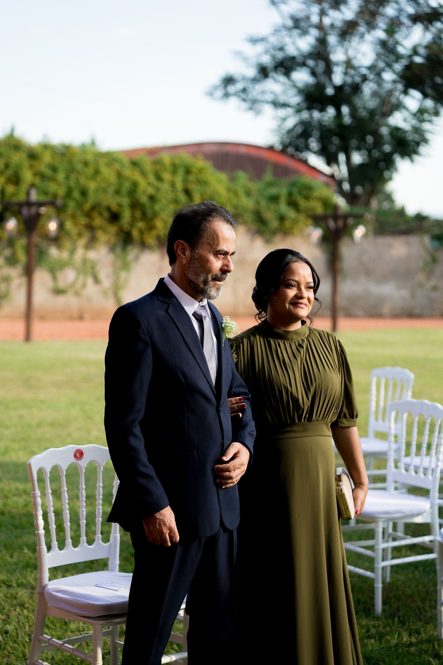 Elegant couple in formal attire walking down outdoor wedding aisle, man in navy suit, woman in olive green dress.