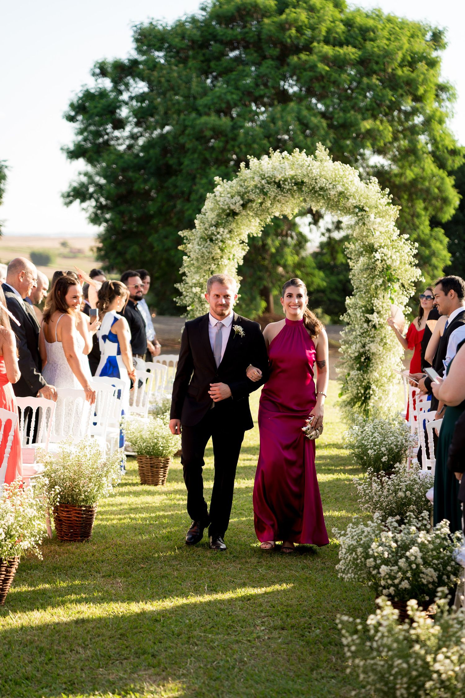 Couple walks down outdoor wedding aisle under floral arch, guests standing, lush green trees backdrop.
