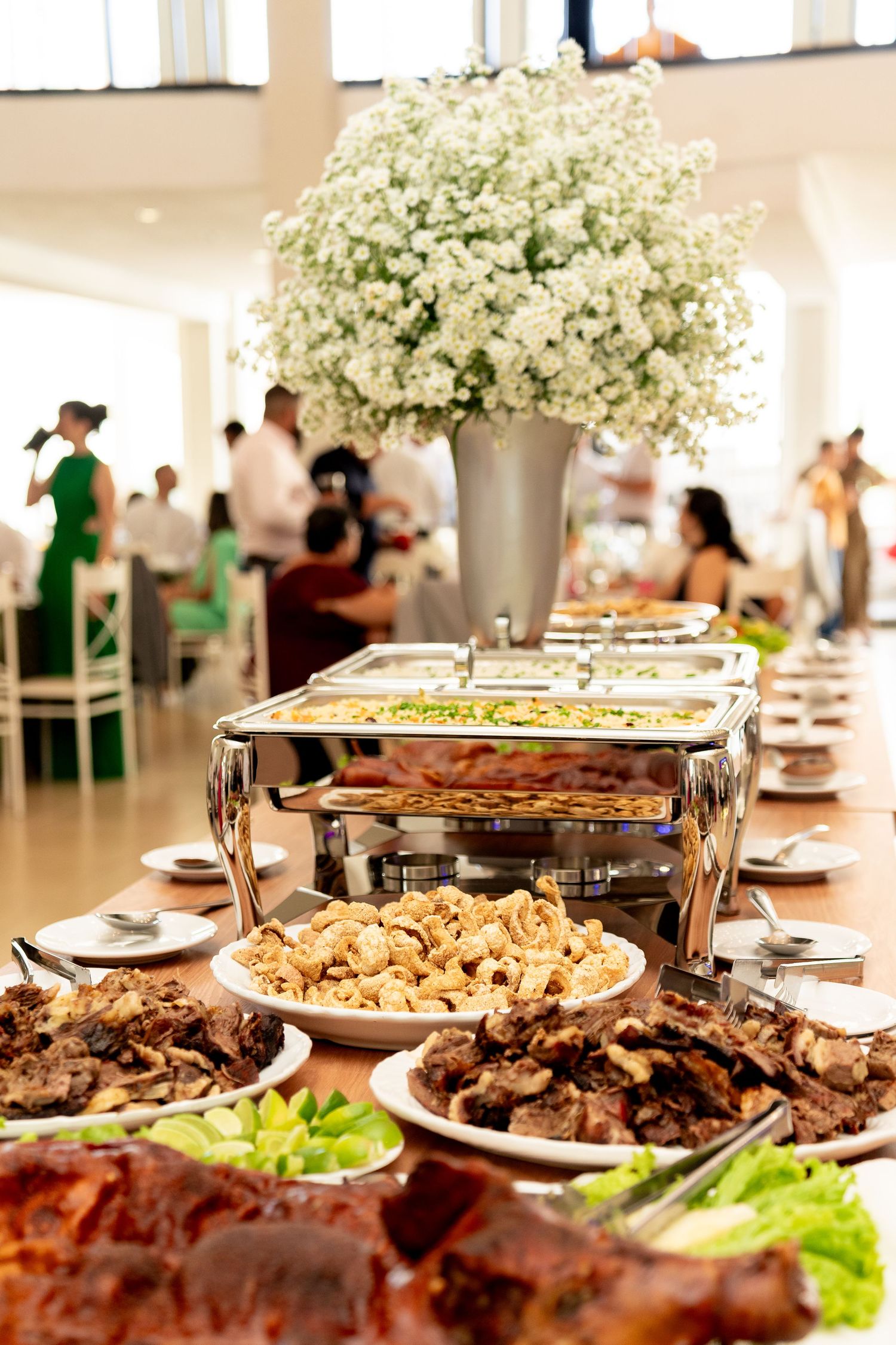 Elegant wedding buffet table with chafing dishes, assorted foods, and a white floral centerpiece in a bright reception hall.