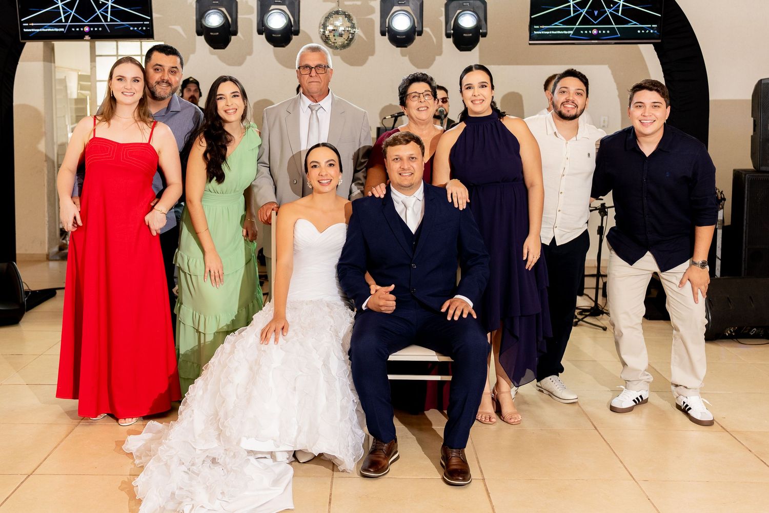 Wedding party group photo with bride in white gown and groom in navy suit surrounded by family members at reception.