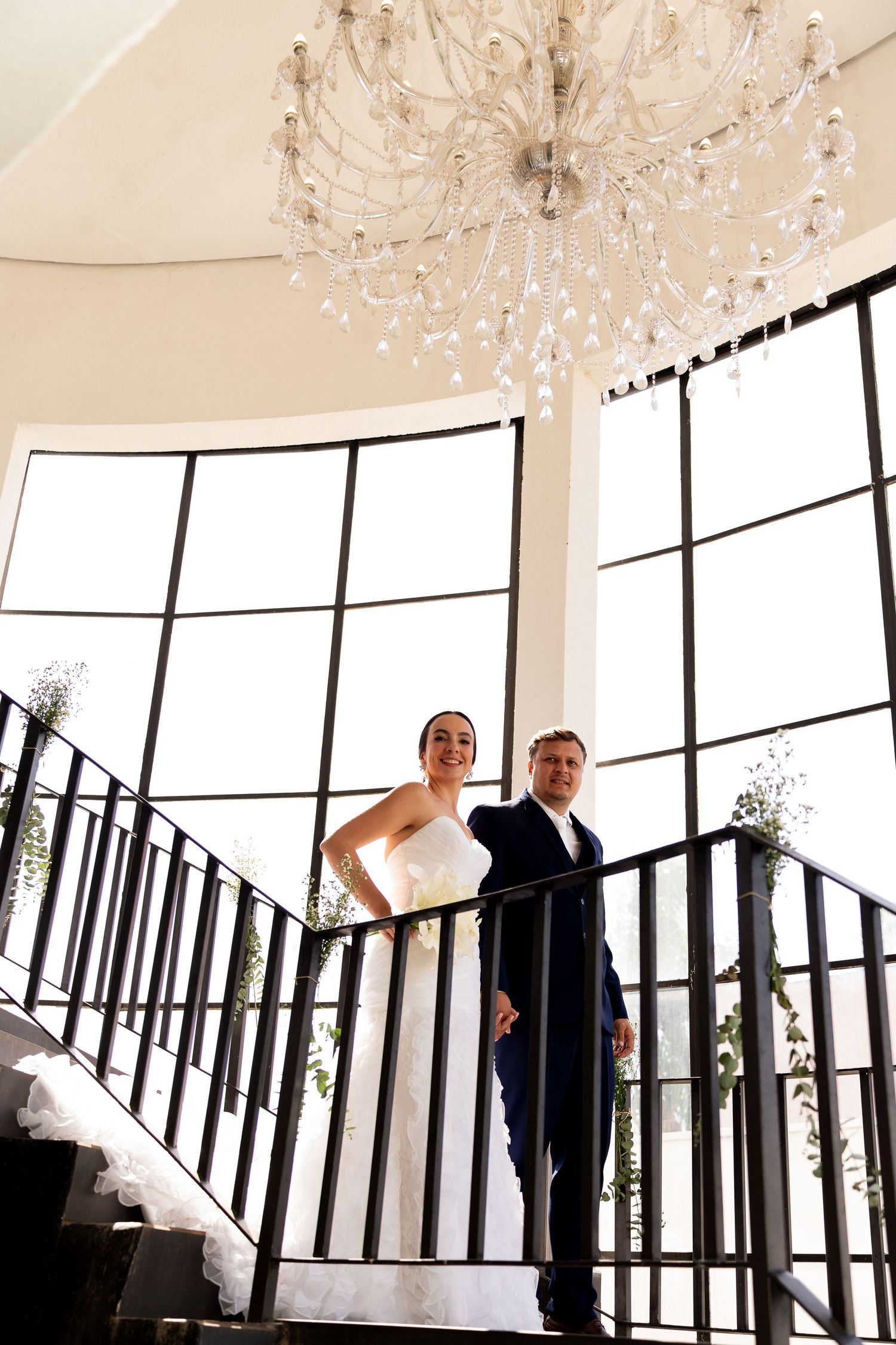Bride and groom standing on elegant staircase beneath crystal chandelier in bright windowed venue.