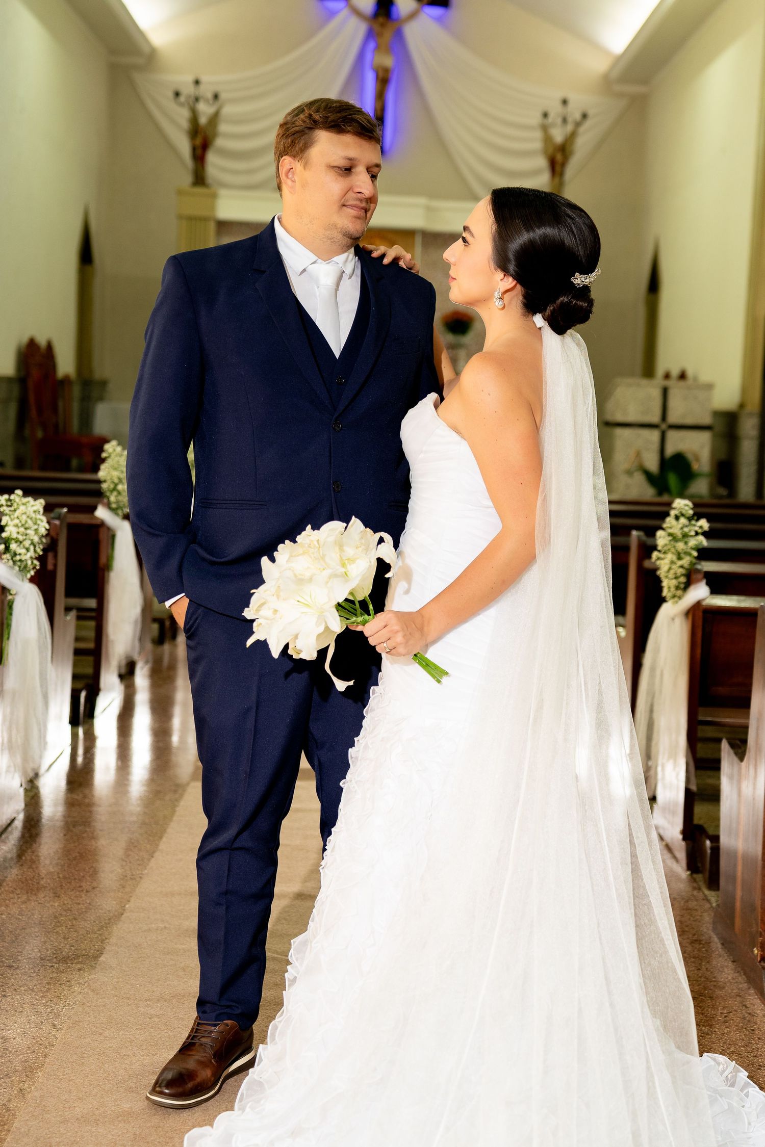 Bride and groom standing together in a church aisle, bride holding white bouquet, groom in navy suit.
