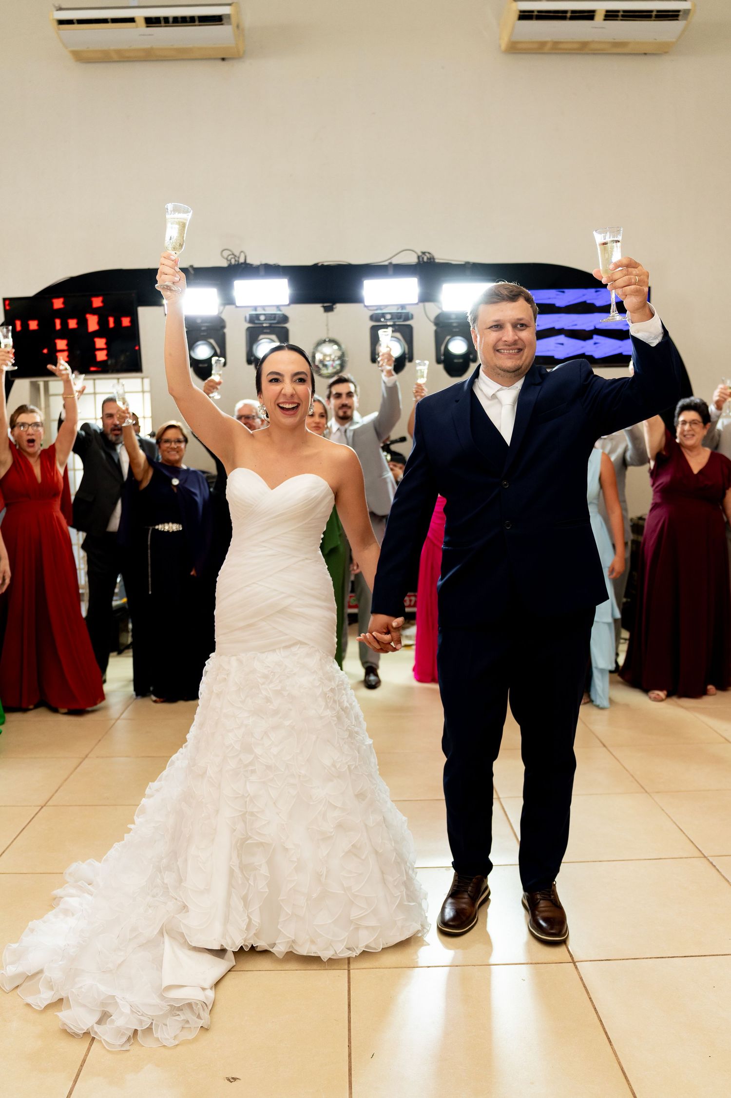 Bride and groom raising champagne glasses at wedding reception with guests celebrating in background.