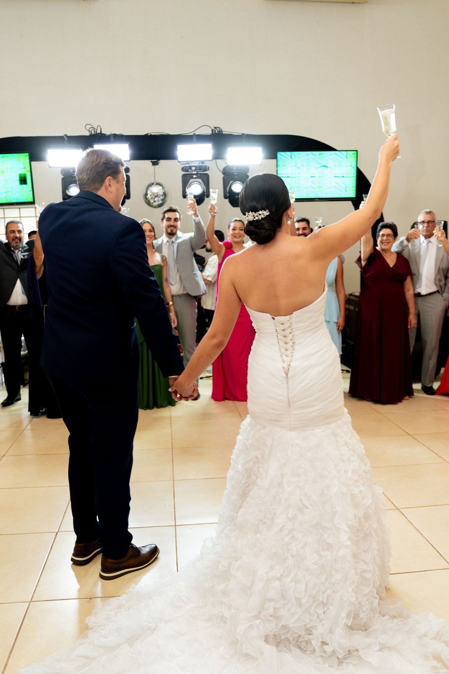 Bride in white lace gown raises champagne glass with groom before wedding guests during reception toast.