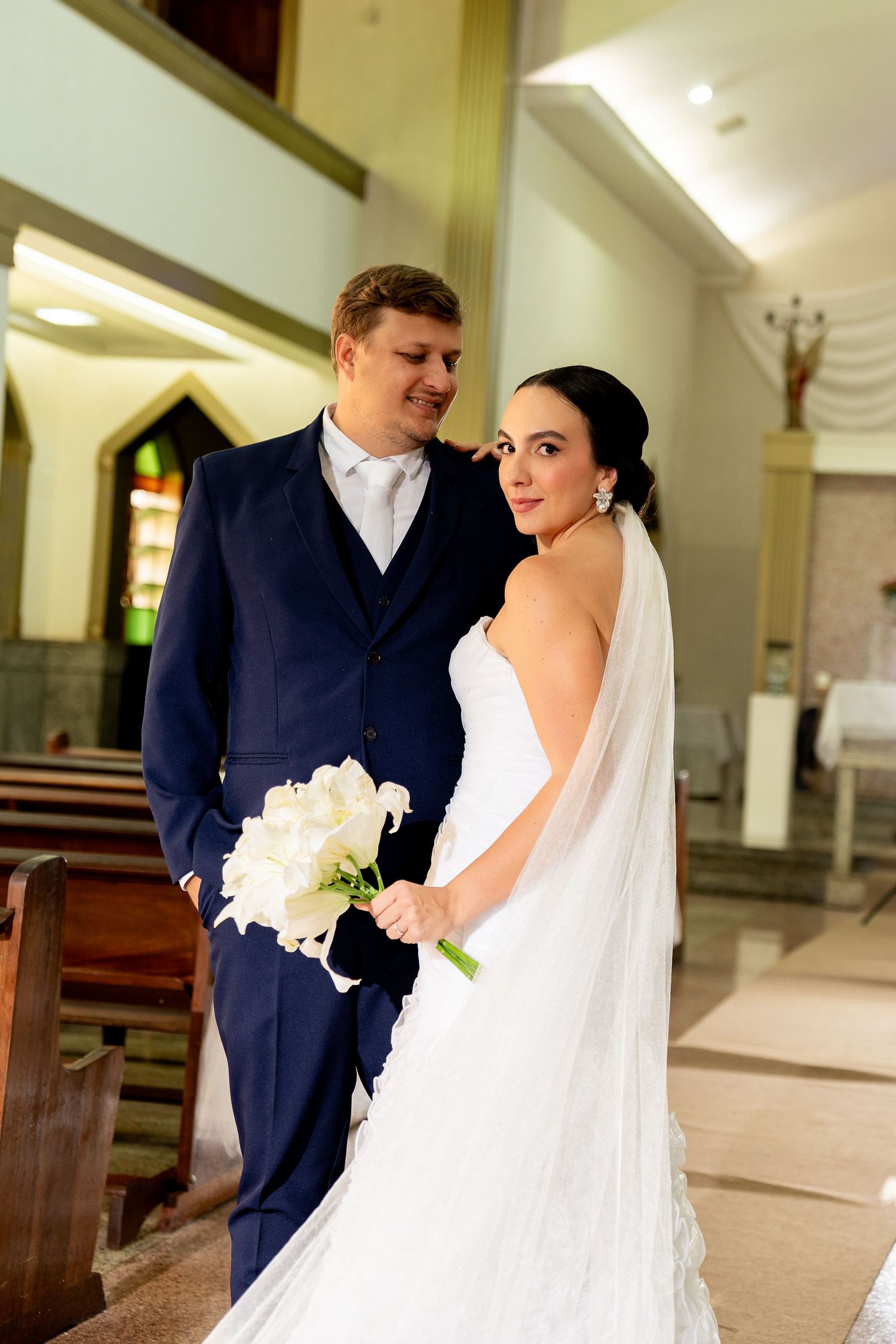 Bride in white gown holding roses bouquet stands with groom in navy suit inside a church.