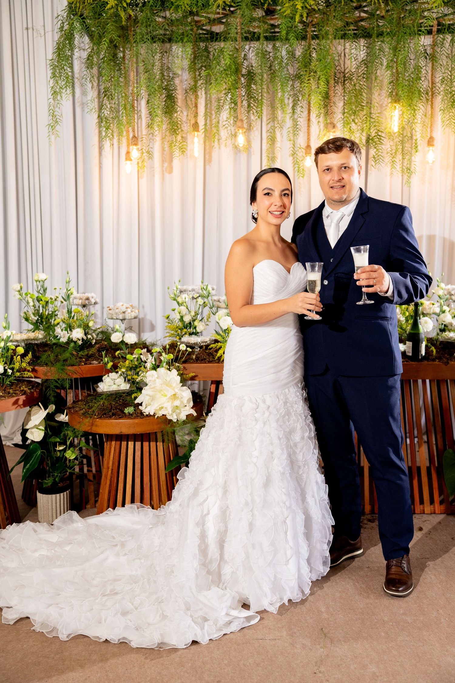 Bride and groom toasting with champagne glasses at elegant wedding reception with floral decor and greenery backdrop.