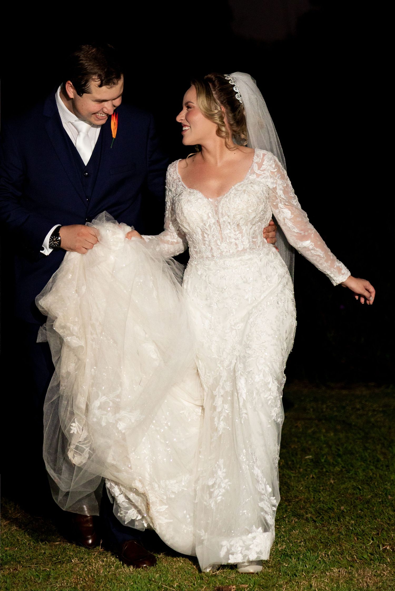 Bride in lace long-sleeve wedding gown and groom in navy suit walk together at night, smiling at each other.