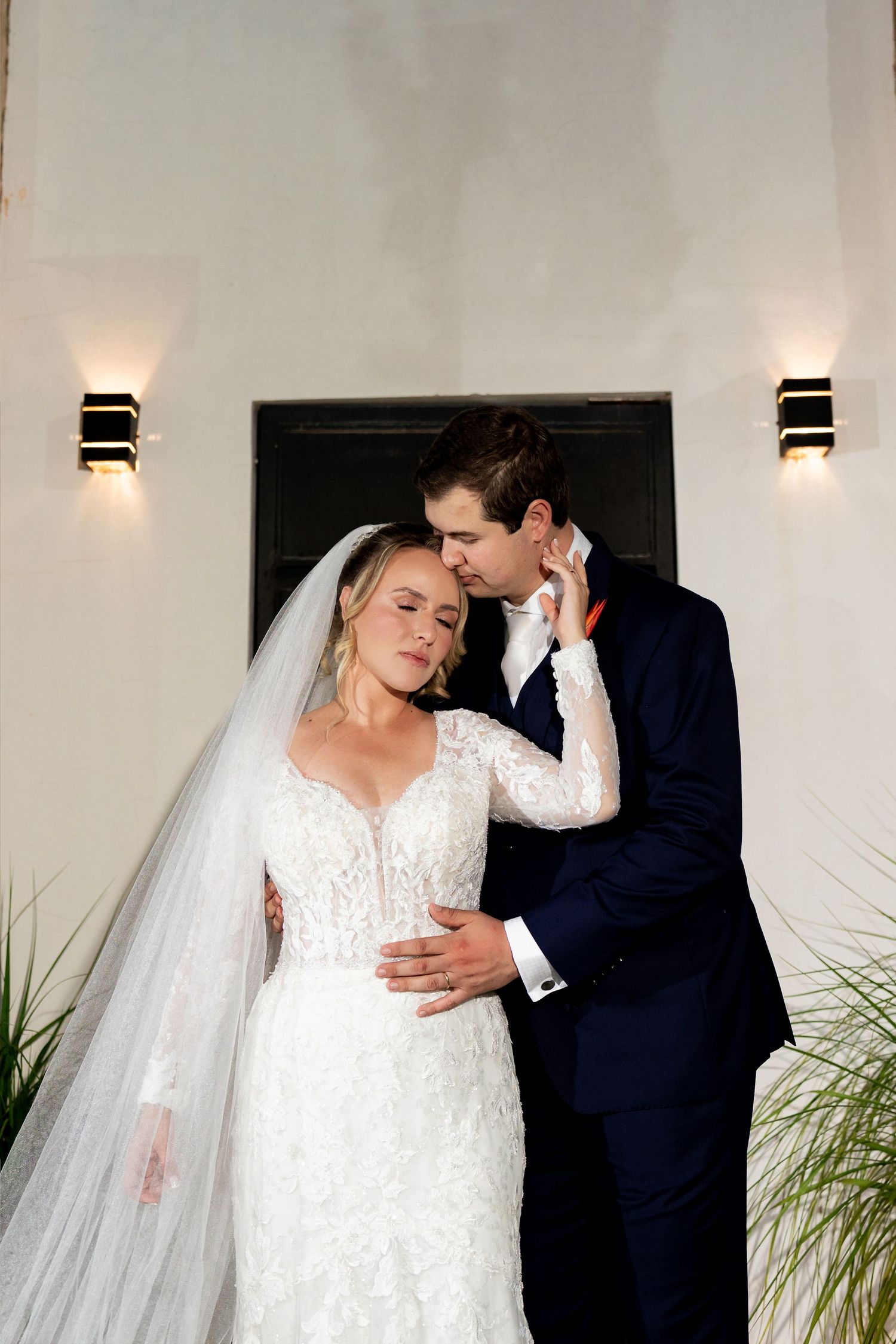Bride in lace gown and veil with groom in navy suit sharing intimate moment by lit wall sconces.