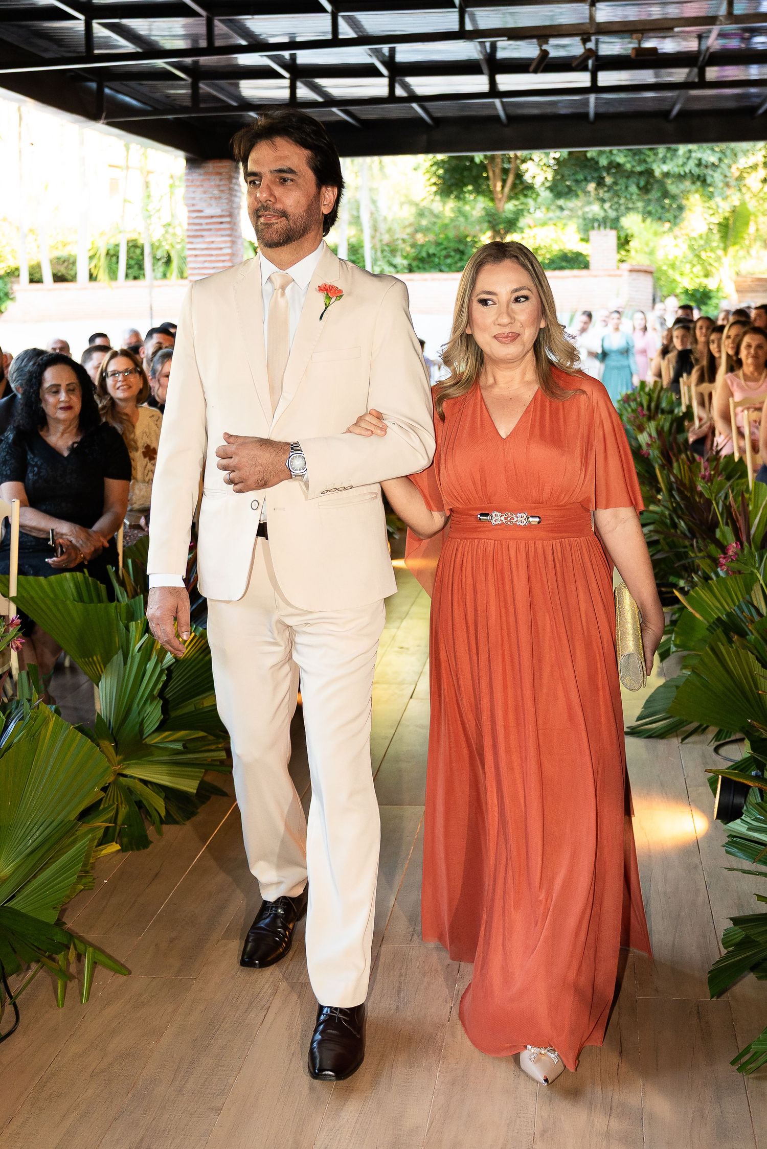 A man in a cream suit and woman in an orange gown walk arm in arm down a wedding aisle lined with tropical plants.