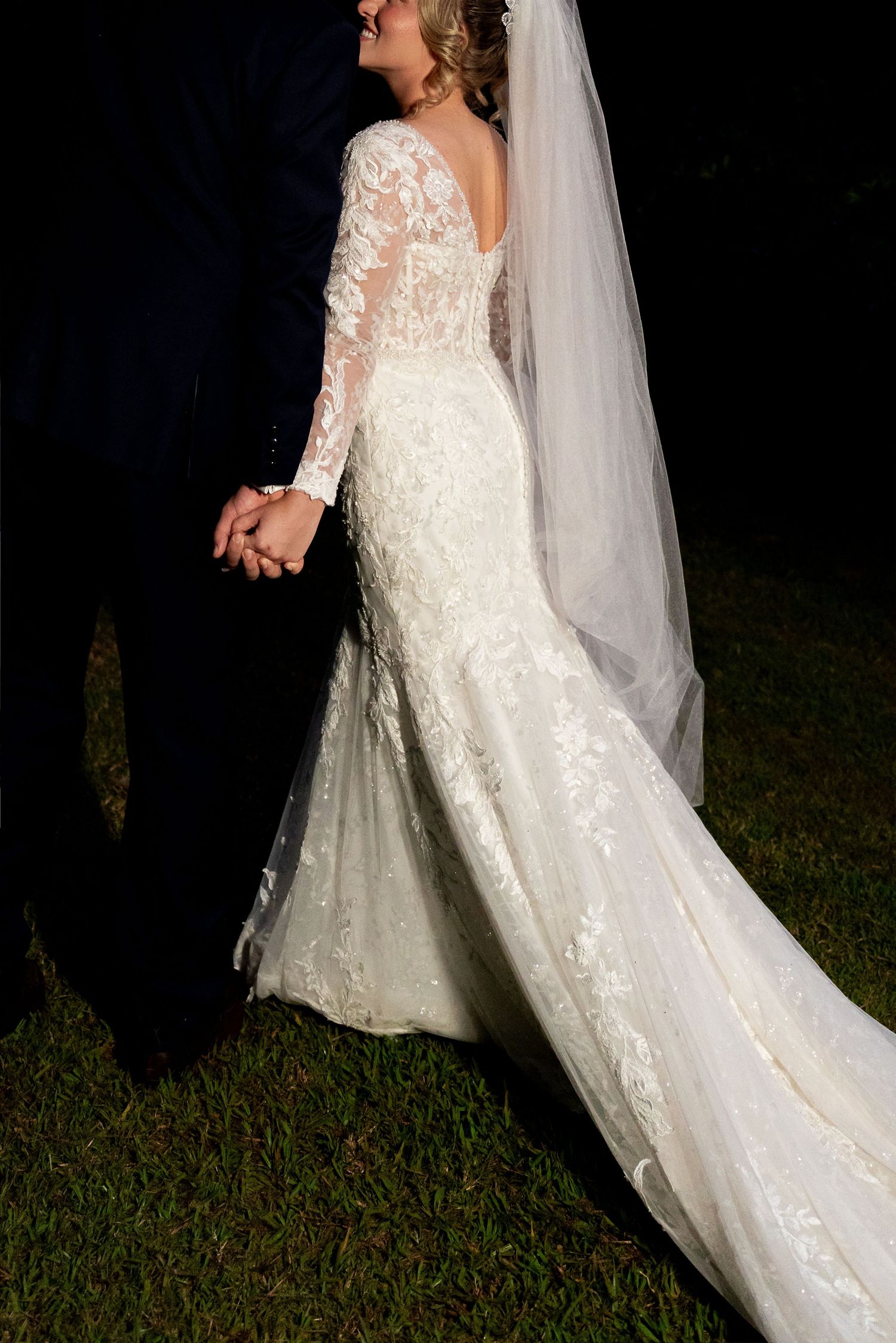 Bride in elegant lace mermaid wedding gown with long sleeves, cathedral veil, holding hands with groom outdoors at night.