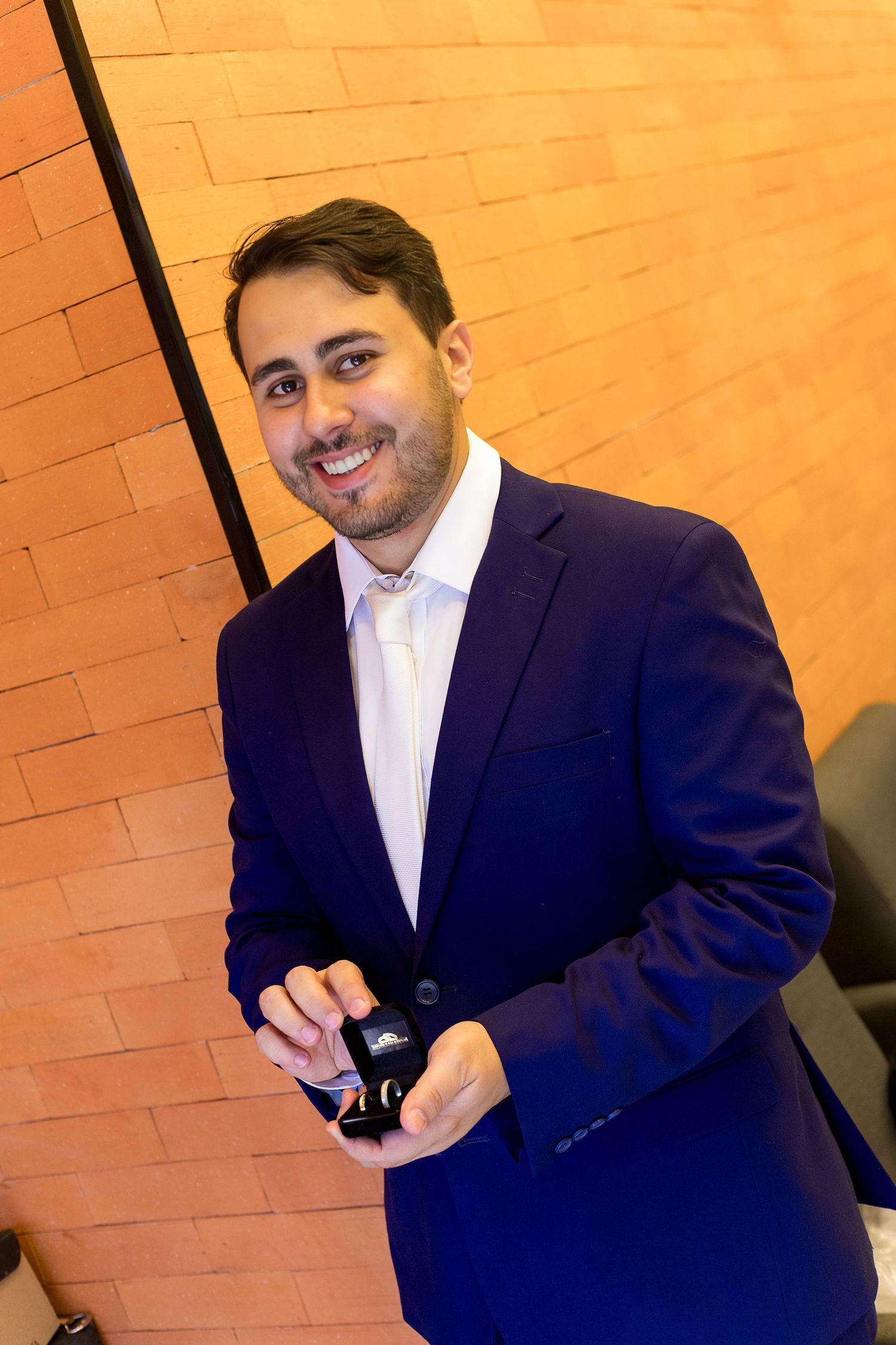 Smiling man in blue blazer holding a small box, standing against an orange brick wall.