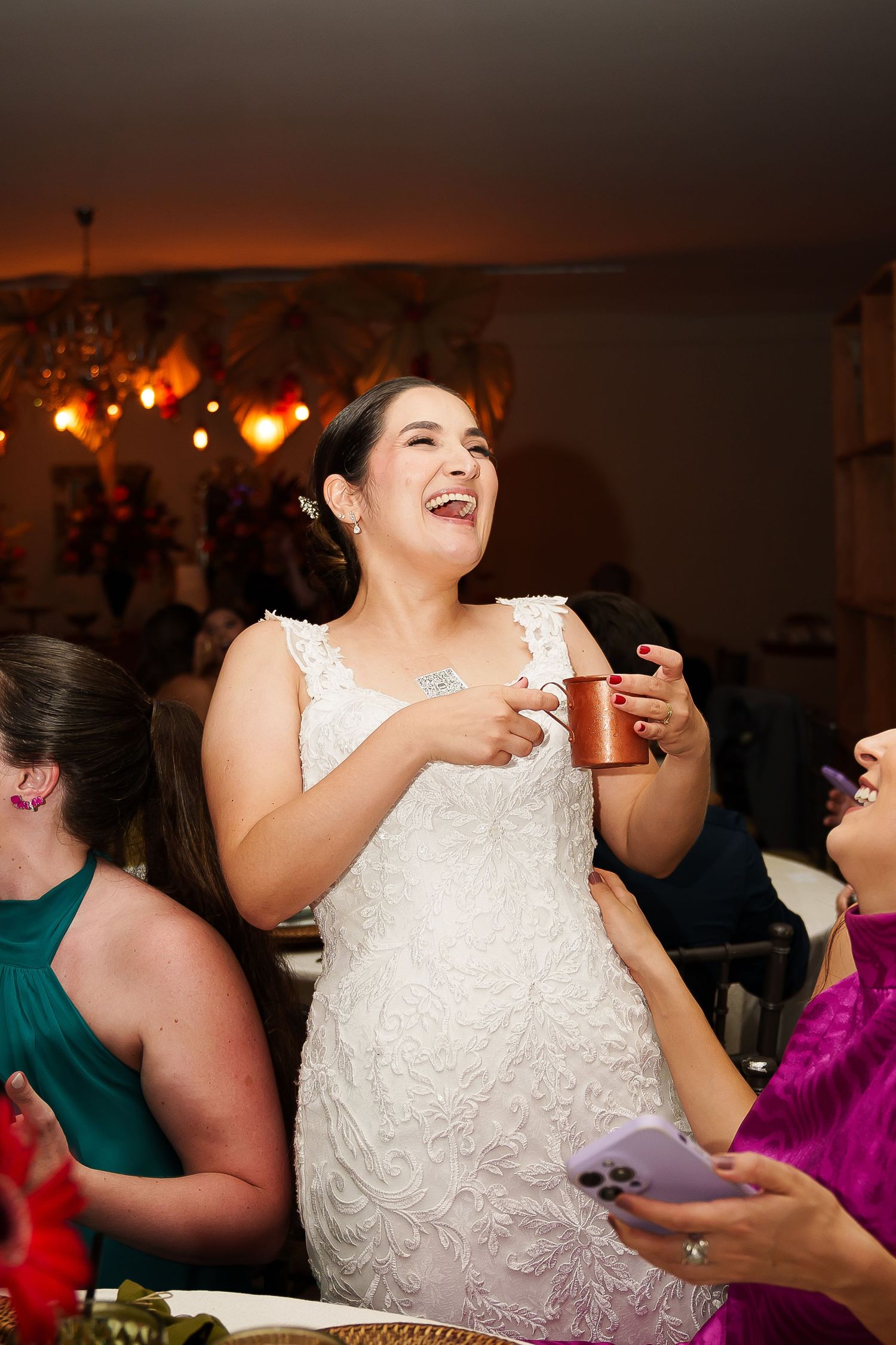Laughing bride in white lace gown holding copper mug at wedding reception surrounded by guests.