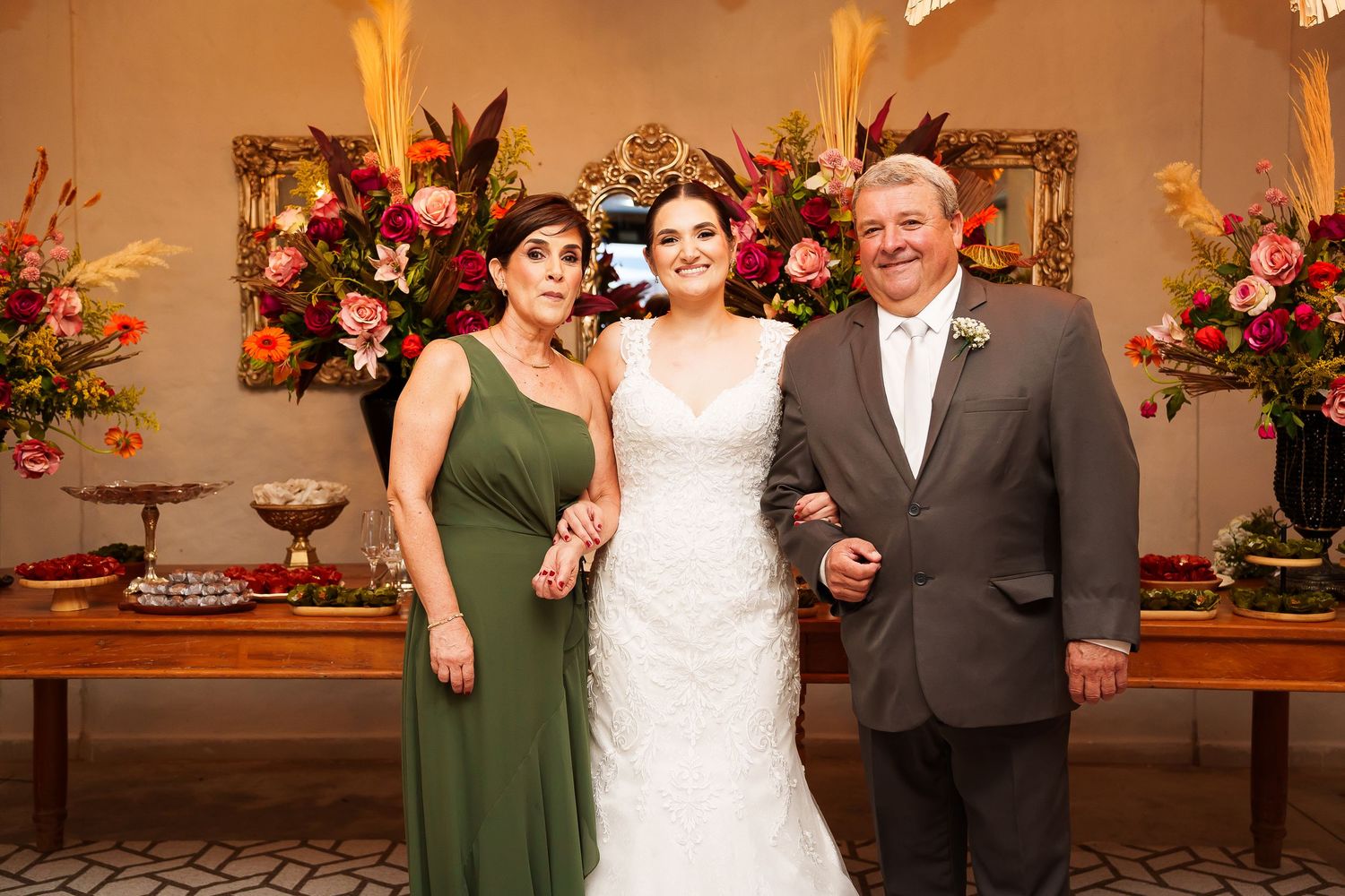 Bride in white gown poses with parents in elegant floral wedding reception setting.