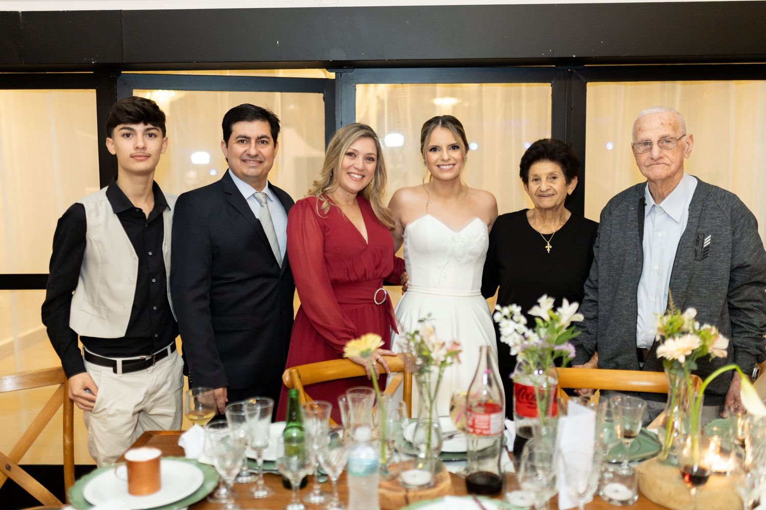 Group of six people posing together at a wedding reception dinner table with floral arrangements.