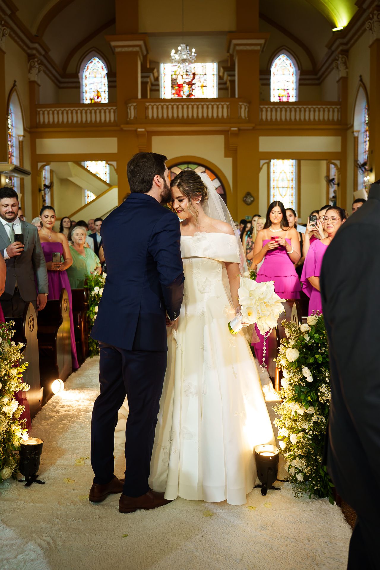 Bride and groom share a kiss at the altar of an elegant church surrounded by candles and wedding guests.