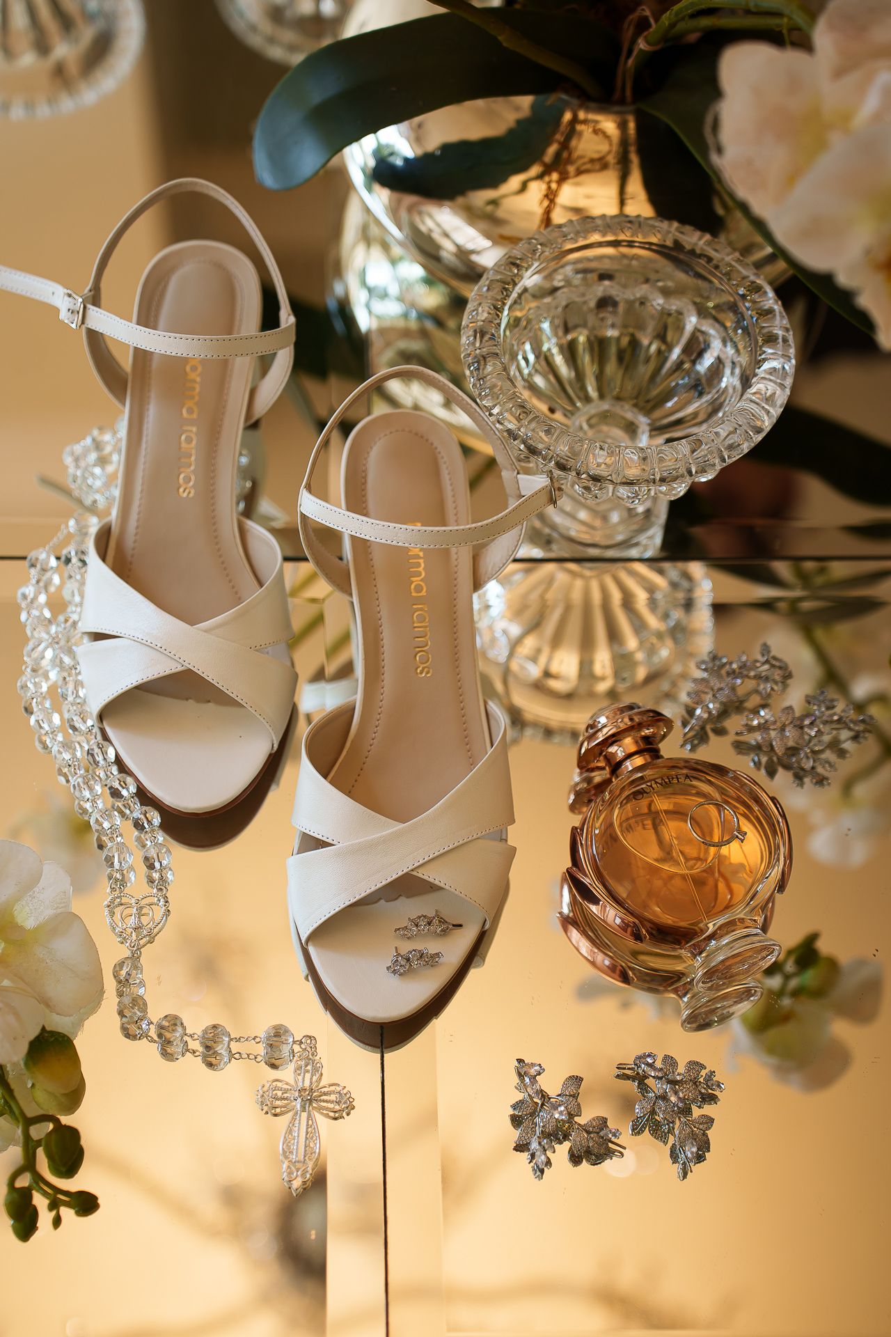 White bridal heels displayed with crystal jewelry, perfume bottle, and floral accents on a mirrored surface.