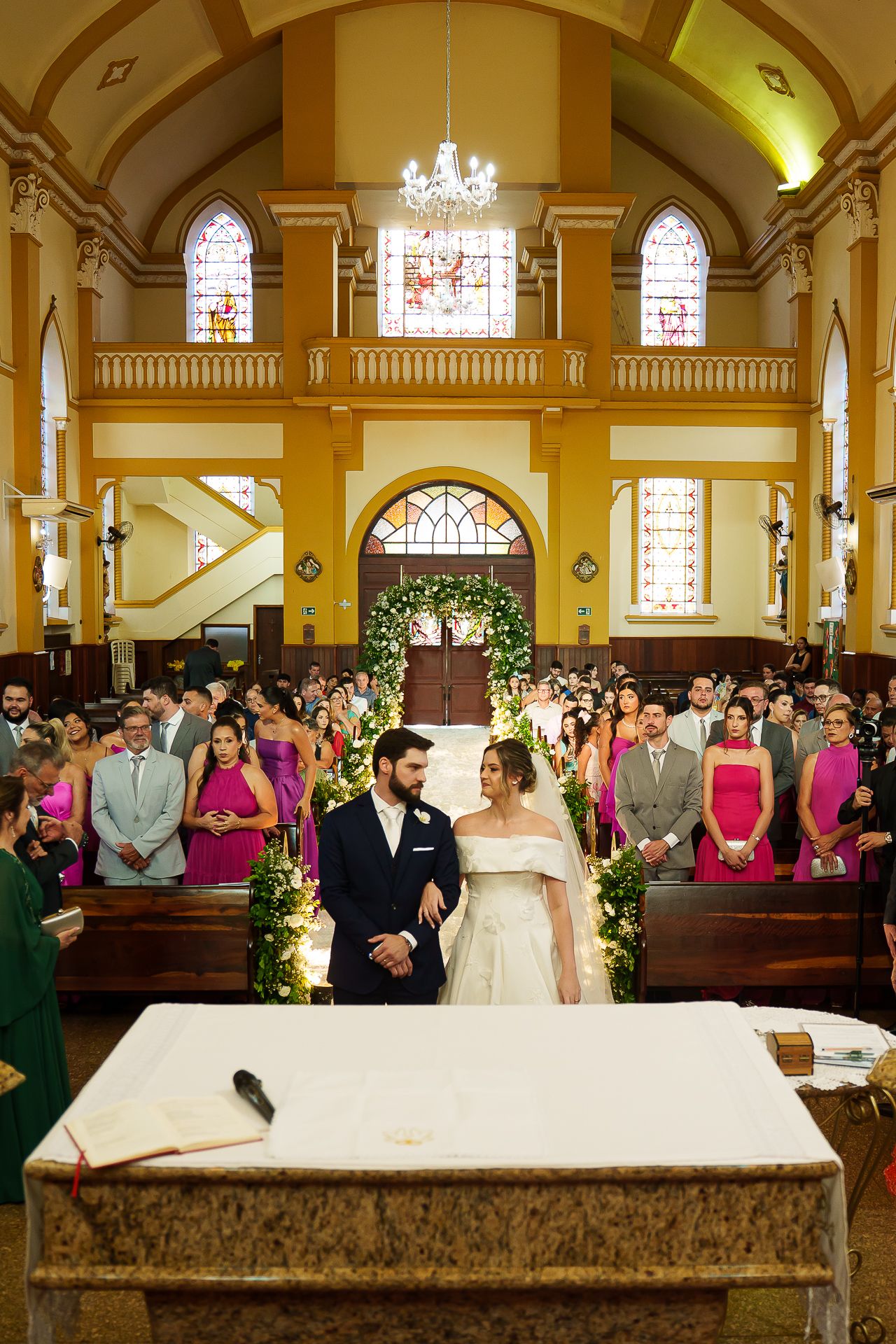 Bride and groom stand at the altar of an elegant church with stained glass windows and pink-dressed bridesmaids lining the aisle.