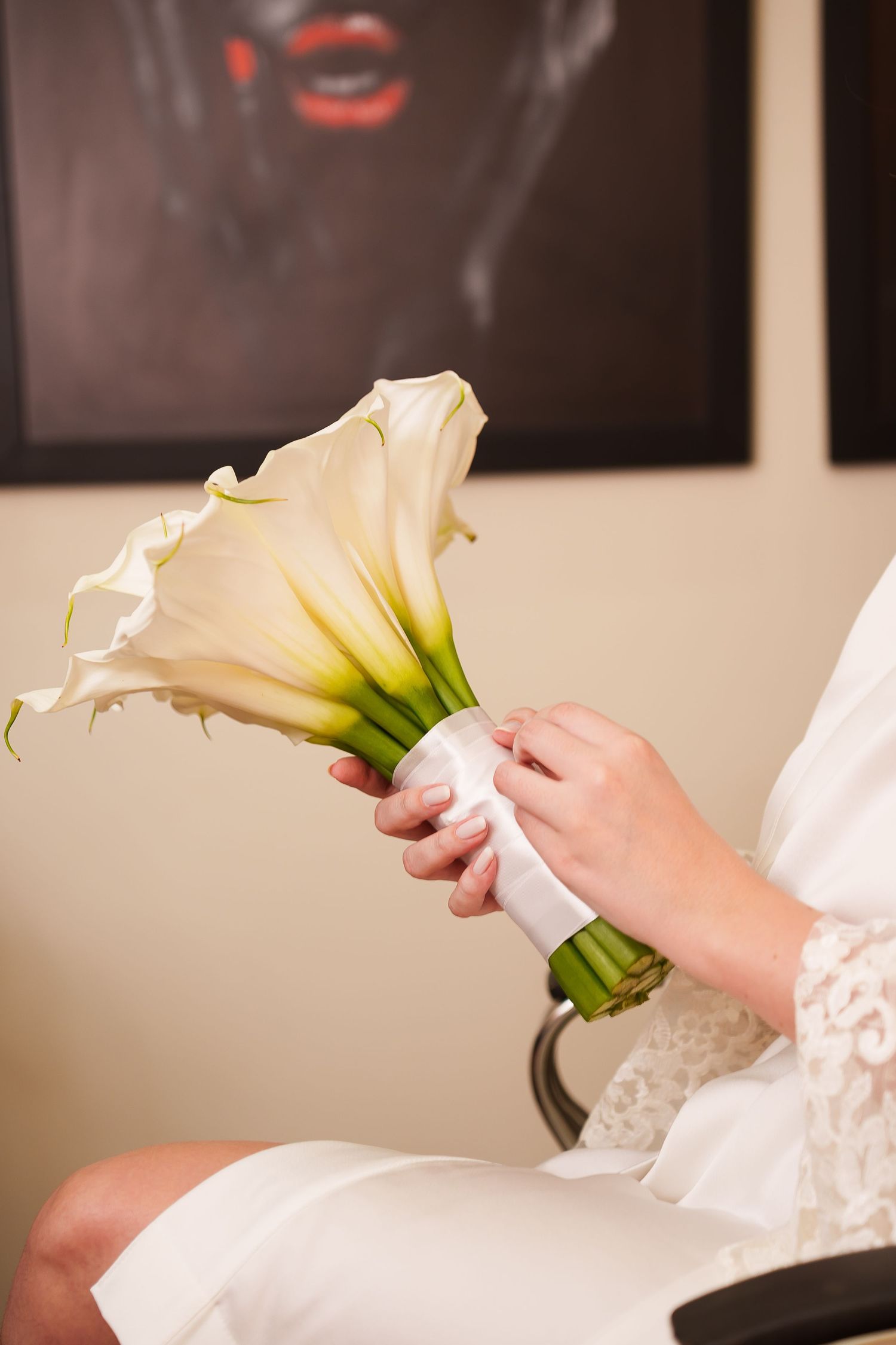 Bride in white robe holding elegant calla lily wedding bouquet wrapped with white ribbon while seated indoors.