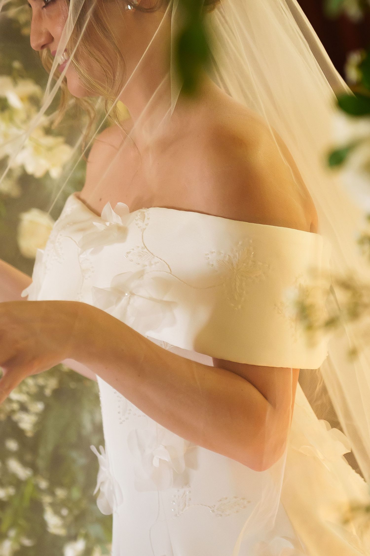 Bride in off-shoulder white wedding gown with floral details and veil, surrounded by soft floral backdrop.
