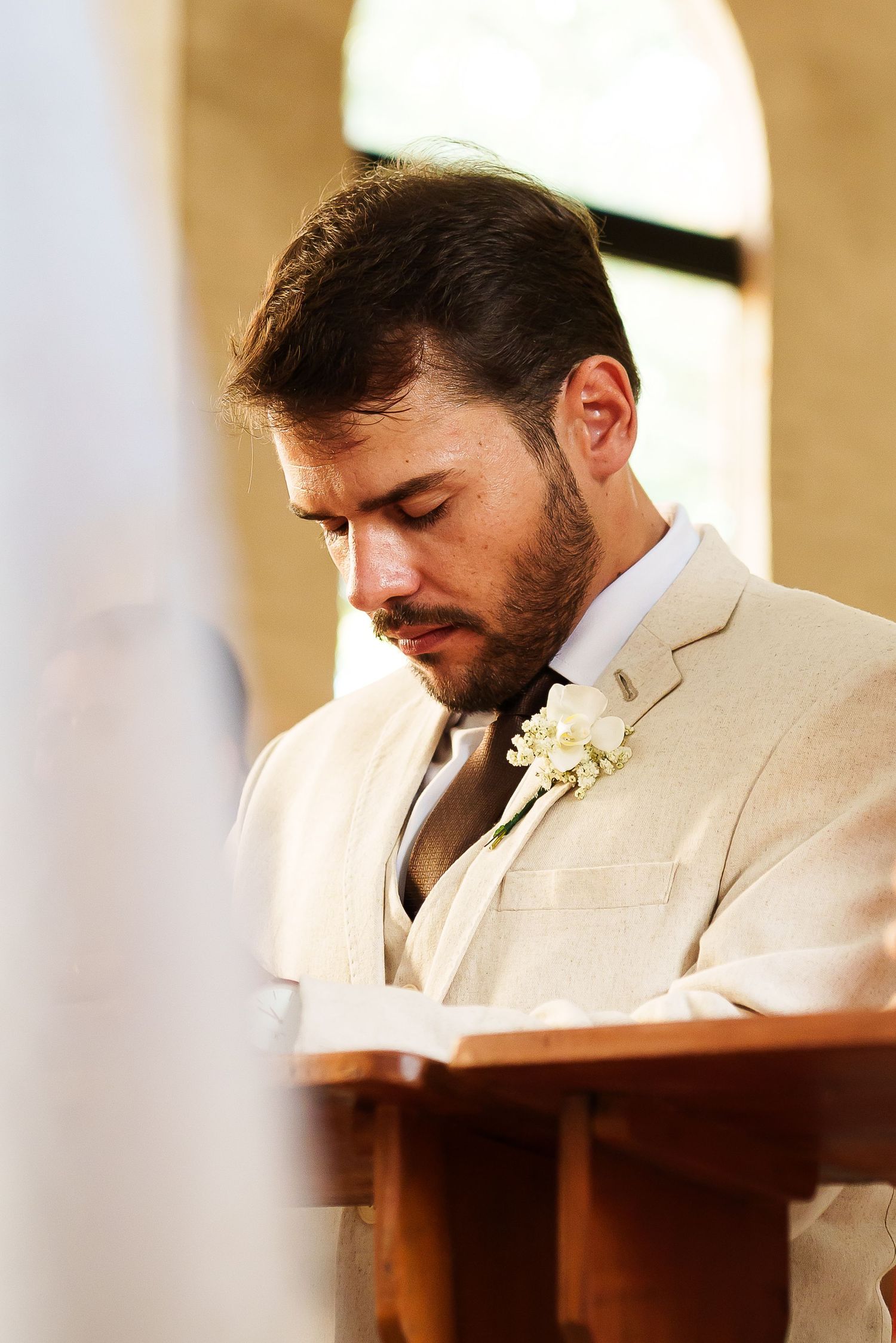 Groom in beige suit with boutonniere bows head in prayer at church altar during wedding ceremony.