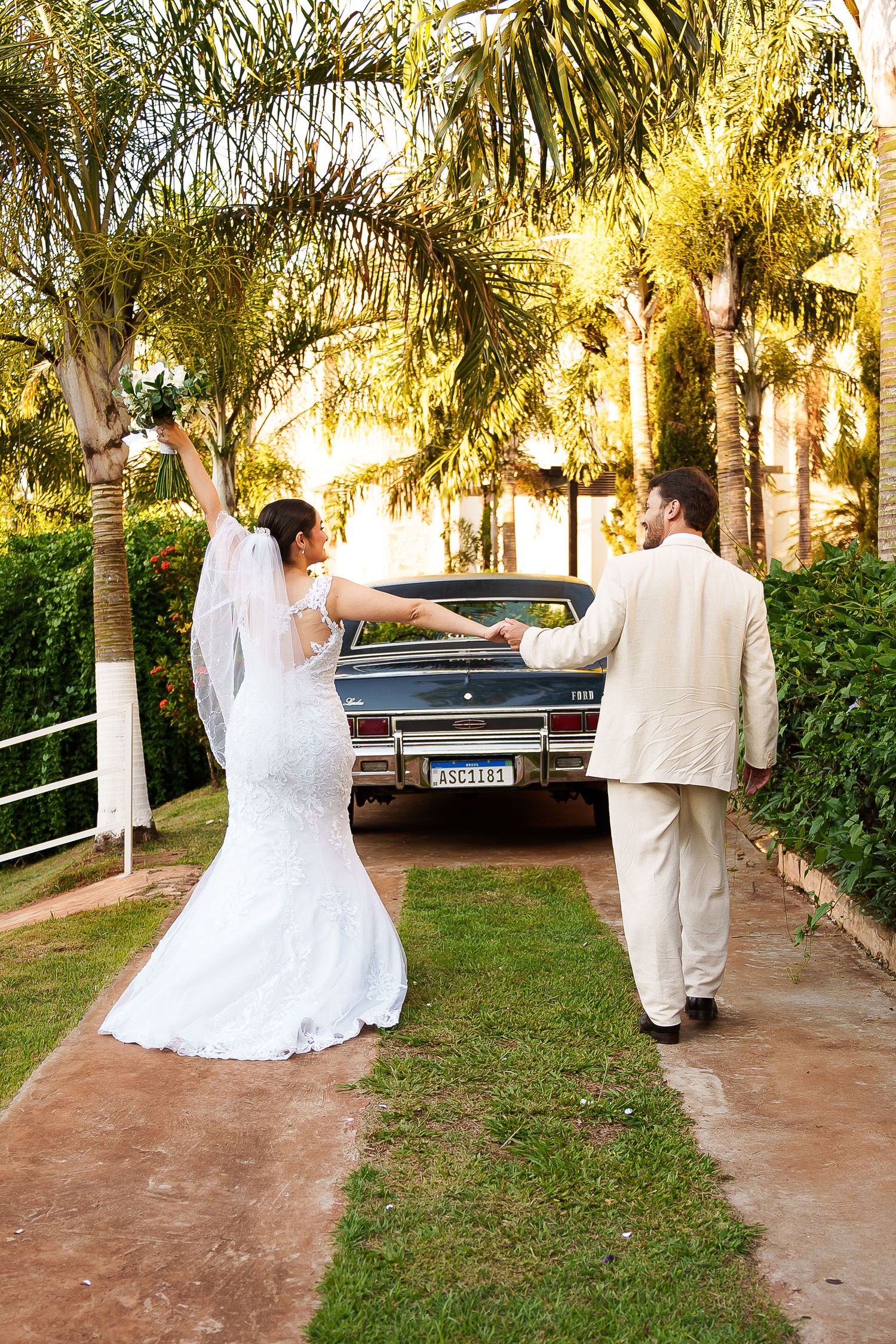 Bride and groom walking hand-in-hand toward a vintage car surrounded by palm trees on their wedding day.