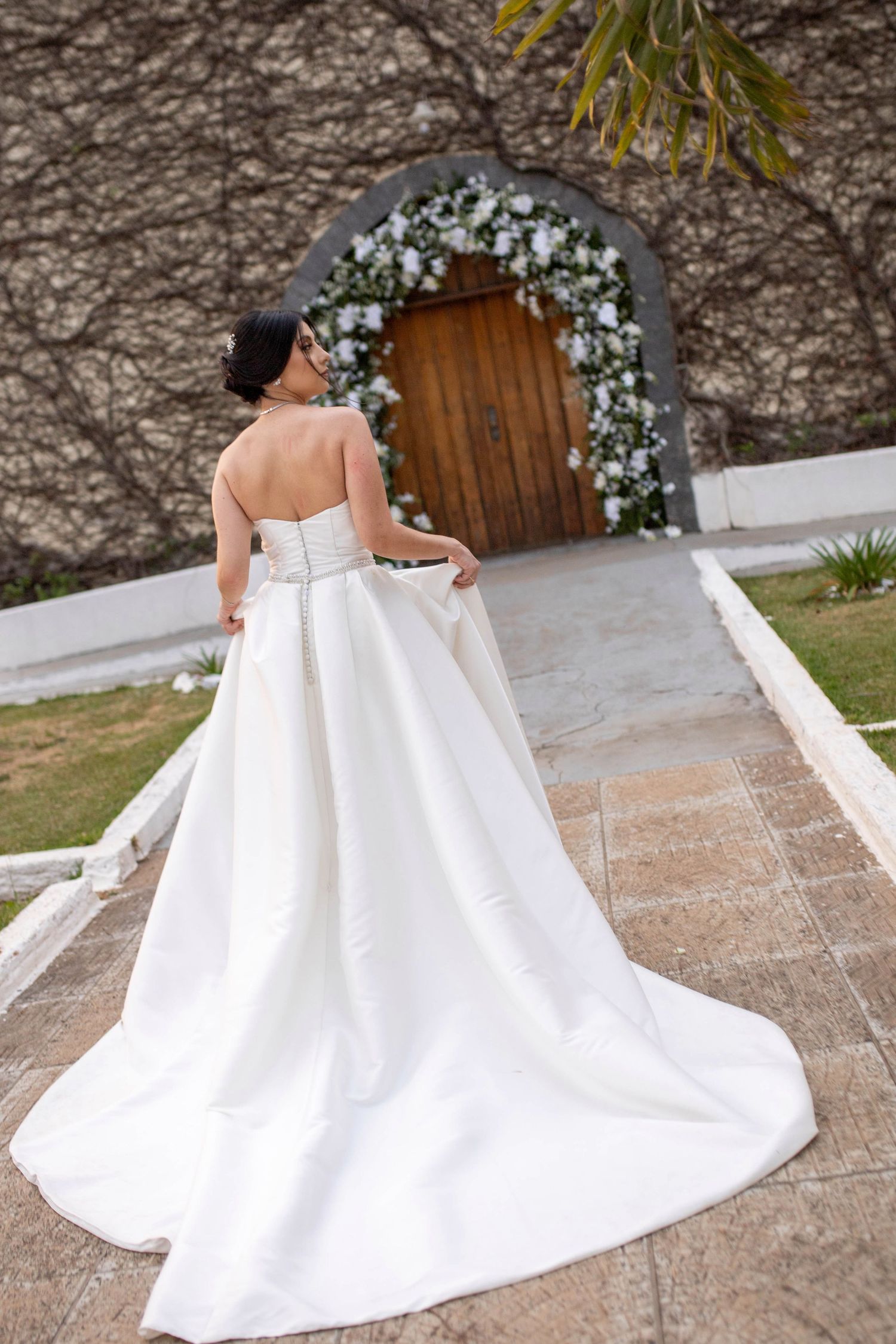 Bride in strapless white gown with long train stands before floral arch and wooden door.