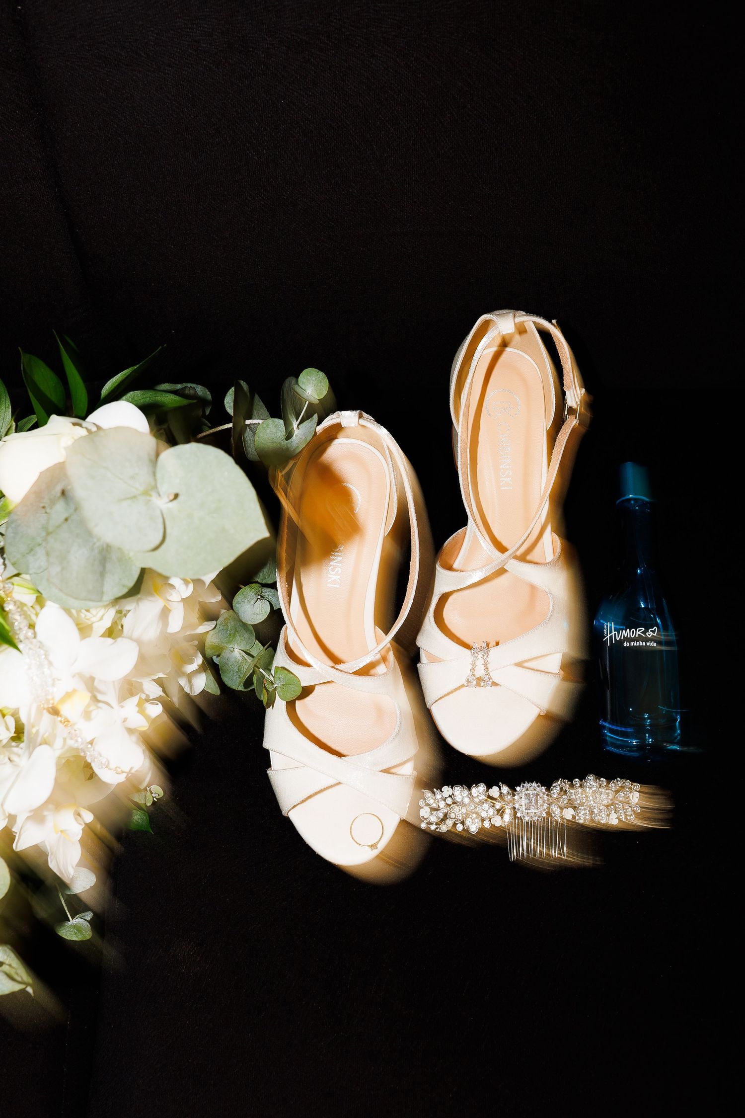 Bridal flat shoes, white floral bouquet, pearl hair comb, and blue perfume bottle arranged on dark background.