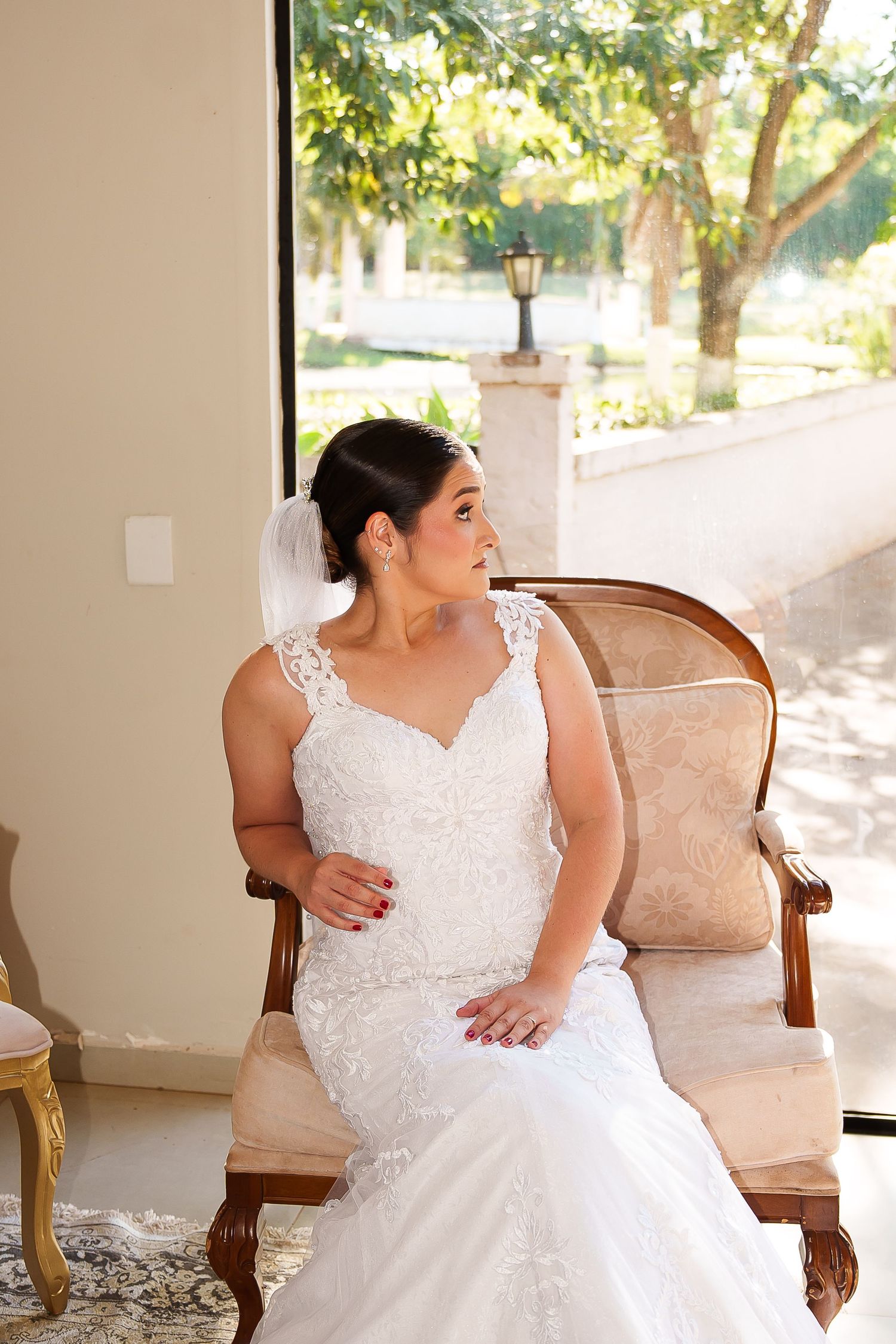 Bride in white lace mermaid gown and veil sits gracefully on vintage chair, gazing toward sunlit garden window.