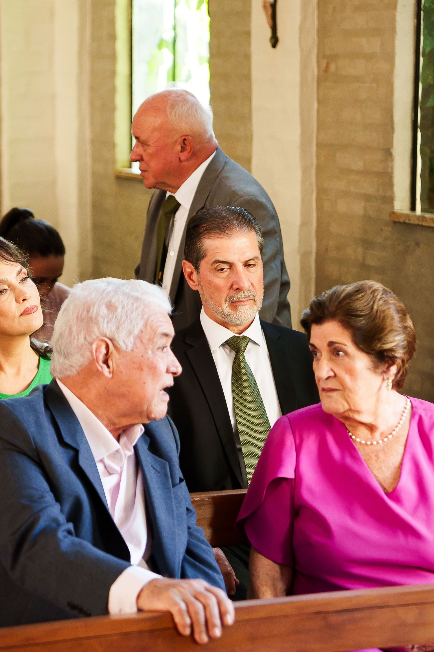 Elderly couple conversing in church pew, surrounded by other well-dressed attendees during a formal religious gathering.